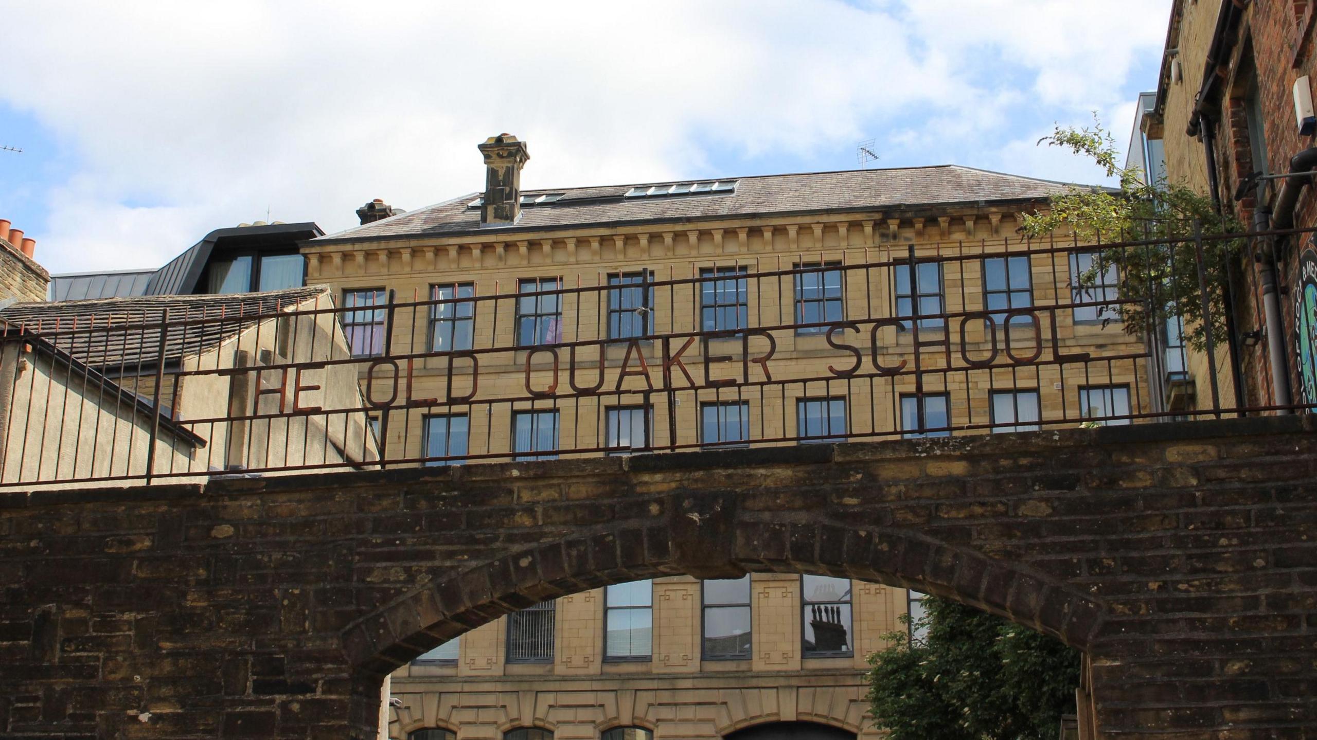 A historic stone archway connects two old brick buildings, with metal railings on top displaying the words THE OLD QUAKER SCHOOL. Behind the arch, a large multi-story stone building with rows of rectangular windows is visible under a partly cloudy blue sky. The scene features traditional architecture with warm-toned stonework and some greenery near the right side of the arch.