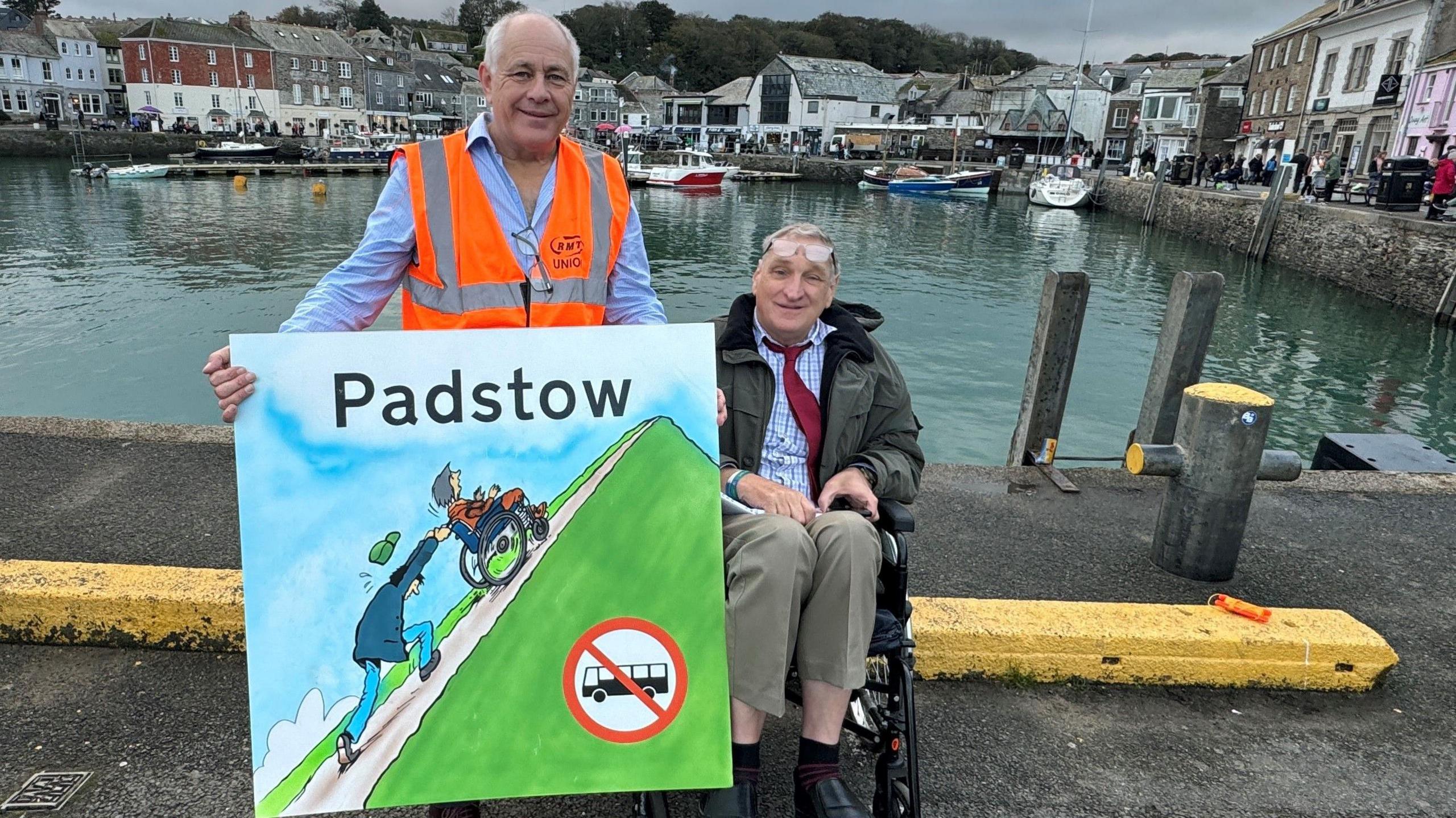 Barry West stands with David Redgwell who sits in a wheelchair beside Padstow harbour. Mr West holds a large sign that shows an illustration of a man struggling to push a person in a wheelchair up a steep hill and there is a 'no buses' sign.
