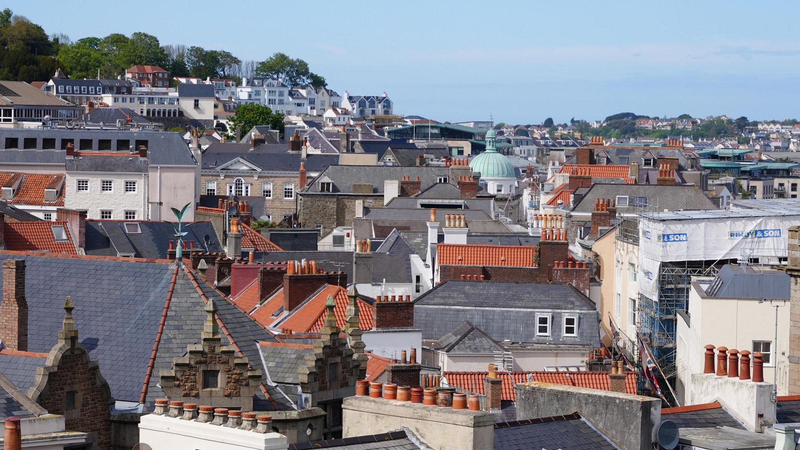 A view looking across the rooftops of St Peter Port. Some of them have terracotta chimney pots and tiles, others are tiled with grey slate. There is a green dome atop a while building in the middle distance. 