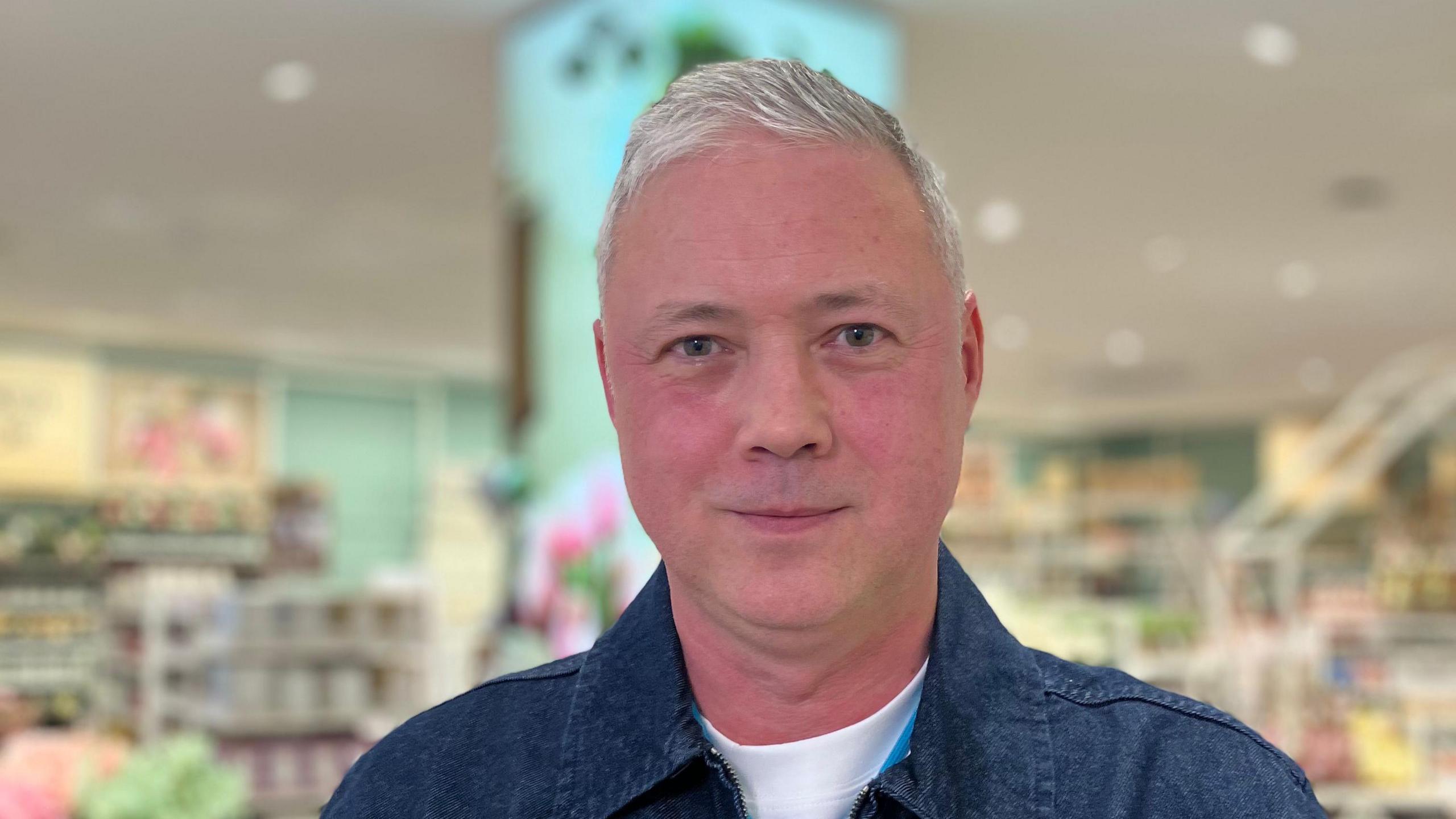 Fintan Costello. He has short white hair, blue eyes, wearing a denim shirt and white t-shirt underneath. Primark home decor can be seen in the background.