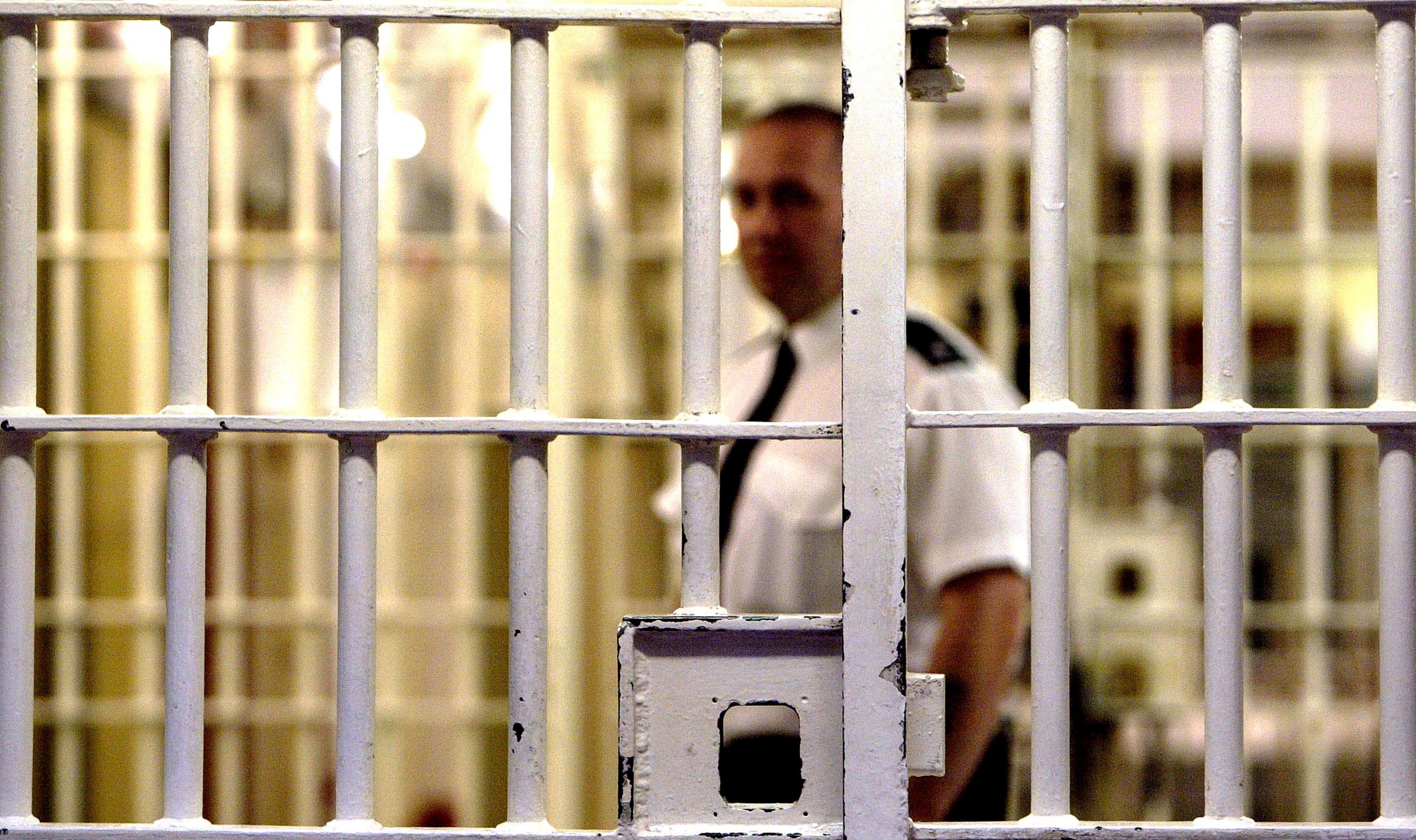 A prison guard stands behind a locked white metal gate inside HMP Pentonville. The gate is in focus with the guard and background blurred. The guard is dark trousers, a white short-sleeved shift and a black tie.