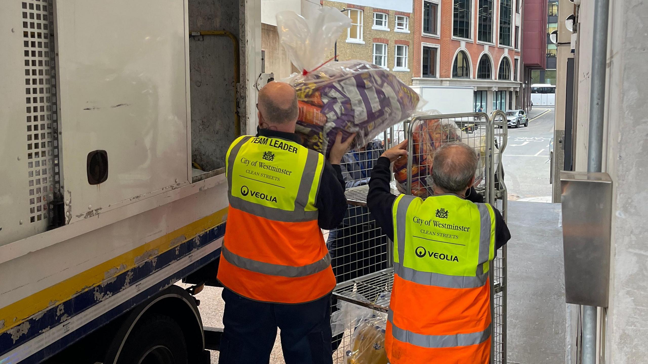 Two men with their backs to the camera wearing high visibility vests in yellow and orange lift clear plastic bags of products off a metal trolley and into a van.