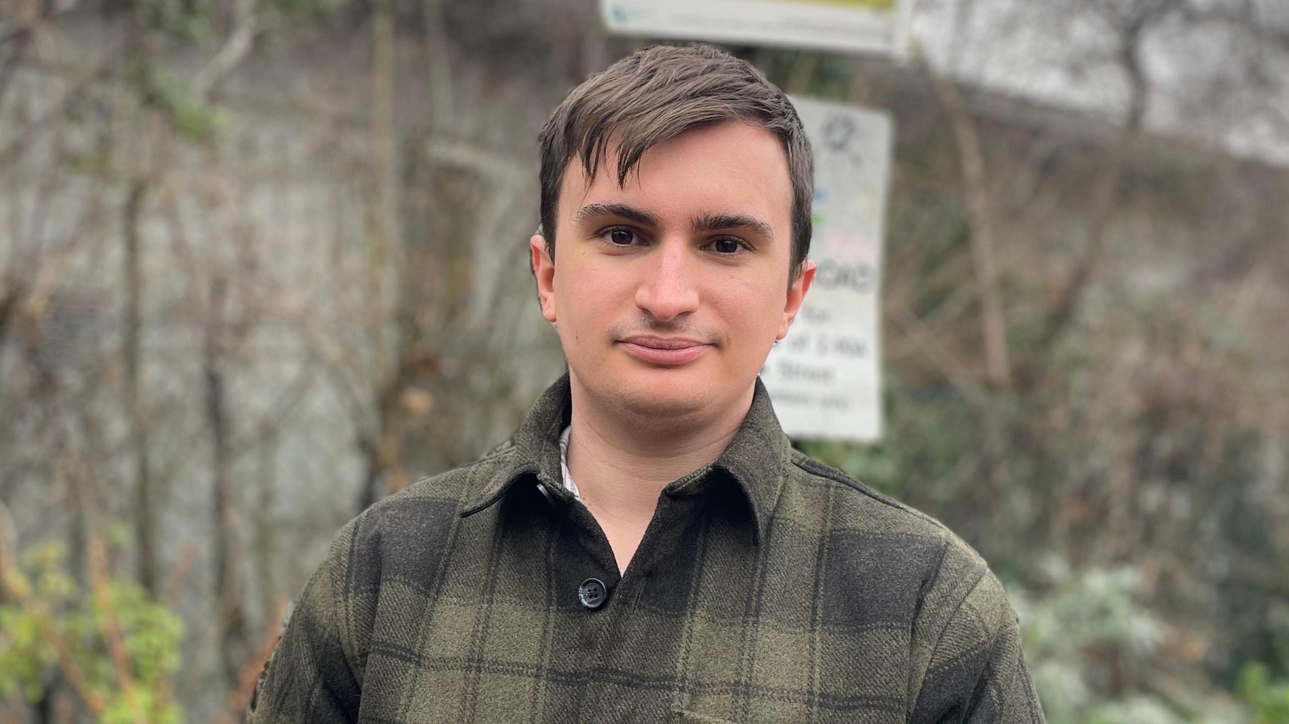 Elliot Tong is looking at the camera, wearing a green checked shirt. In the background is a tall fence that separates the street from the Beehive Centre.