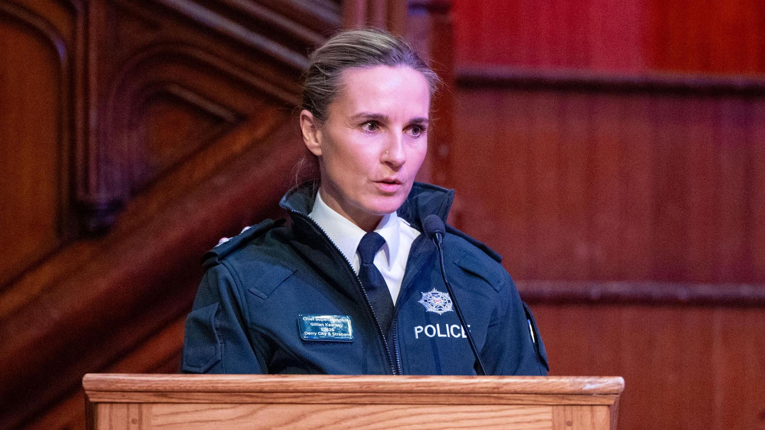 Gillian Kearney is speaking at the pulpit. She has a police uniform on, blonde hair that is pulled back and a white shirt and tie on. 