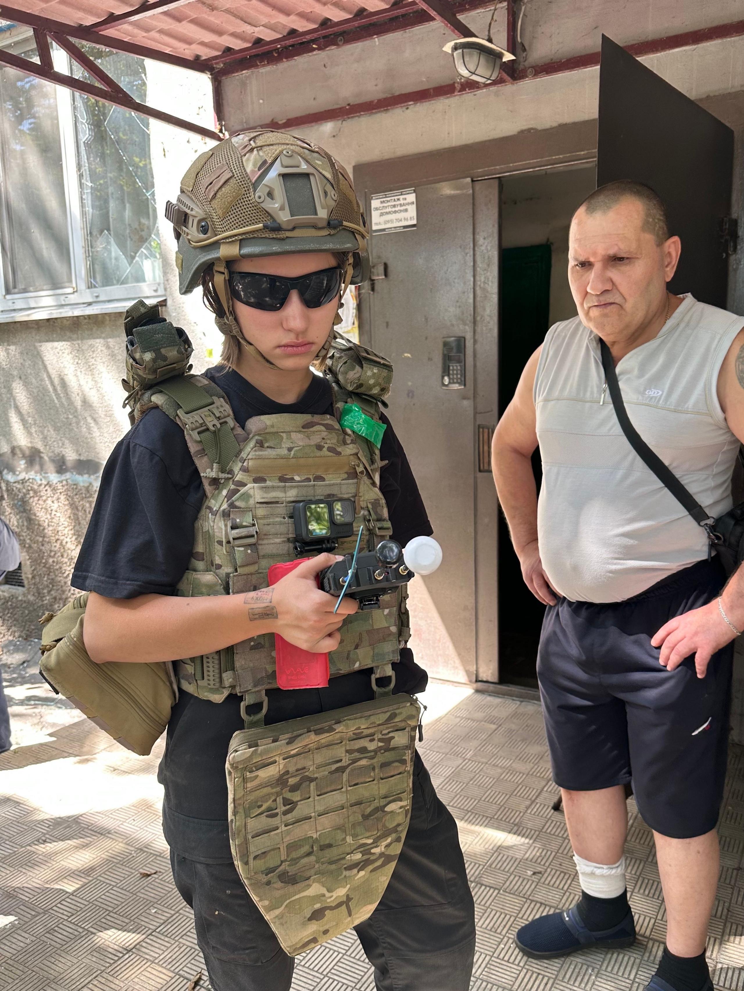 Young woman Varia wearing khaki camouflage gear holding a device, standing next to a middle-aged man wearing a grey vest and blue trousers