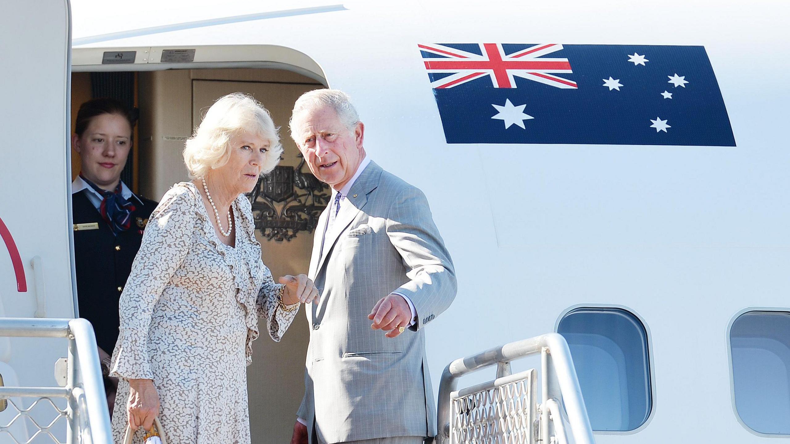 King Charles and Queen Camilla stand at the top of a staircase leading on to a plane, on which a stewardess waits for the pair, next to an Australian flag.