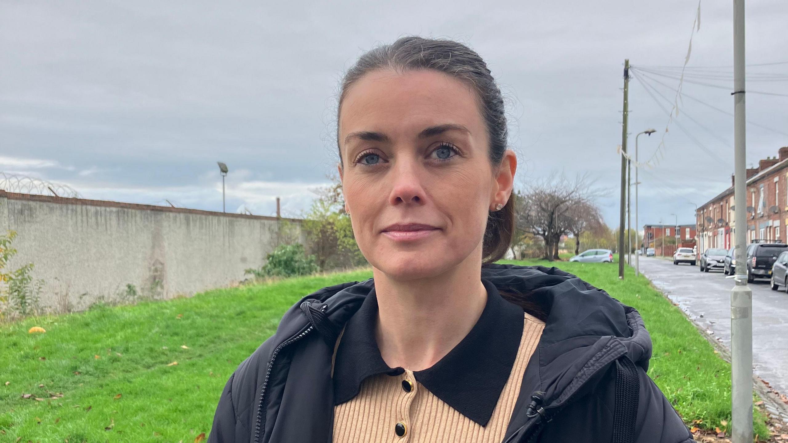 Councillor Lucy Williams stands outside on a roadside in front of a patch of grass. She is wearing a black coat and has brown hair pulled back into a ponytail. 