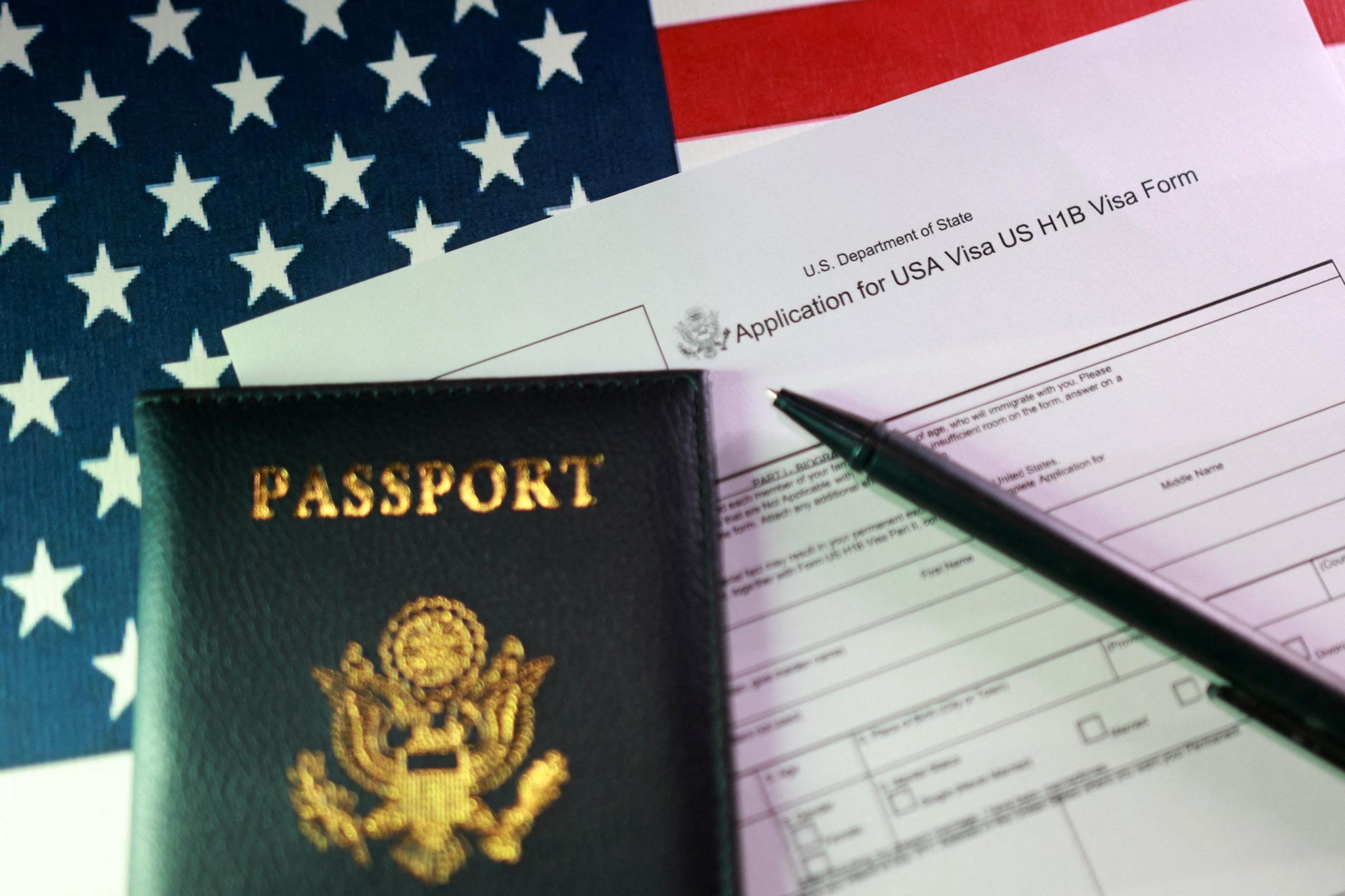 An application for an H-1B visa sits on a table alongside a US passport, a pen and an American flag.