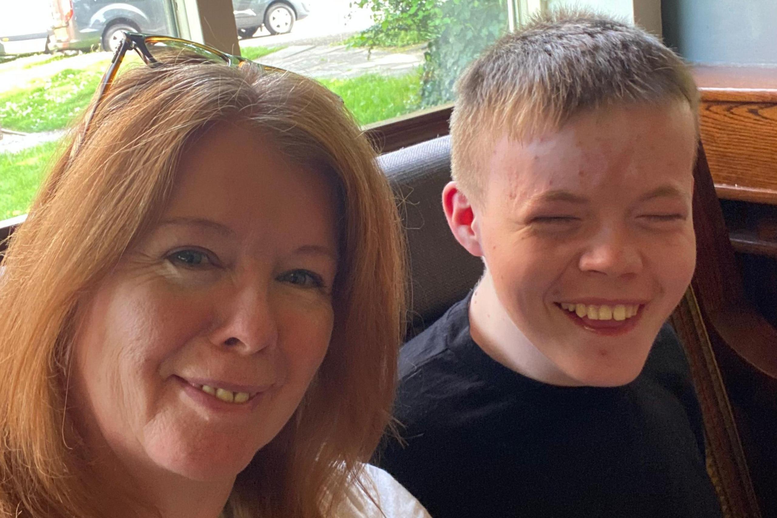 Louise and her son Leo are sitting next to each other. They are both smiling. Leo has short hair and is wearing a black t-shirt and Louise has a pair of glasses resting on her head.