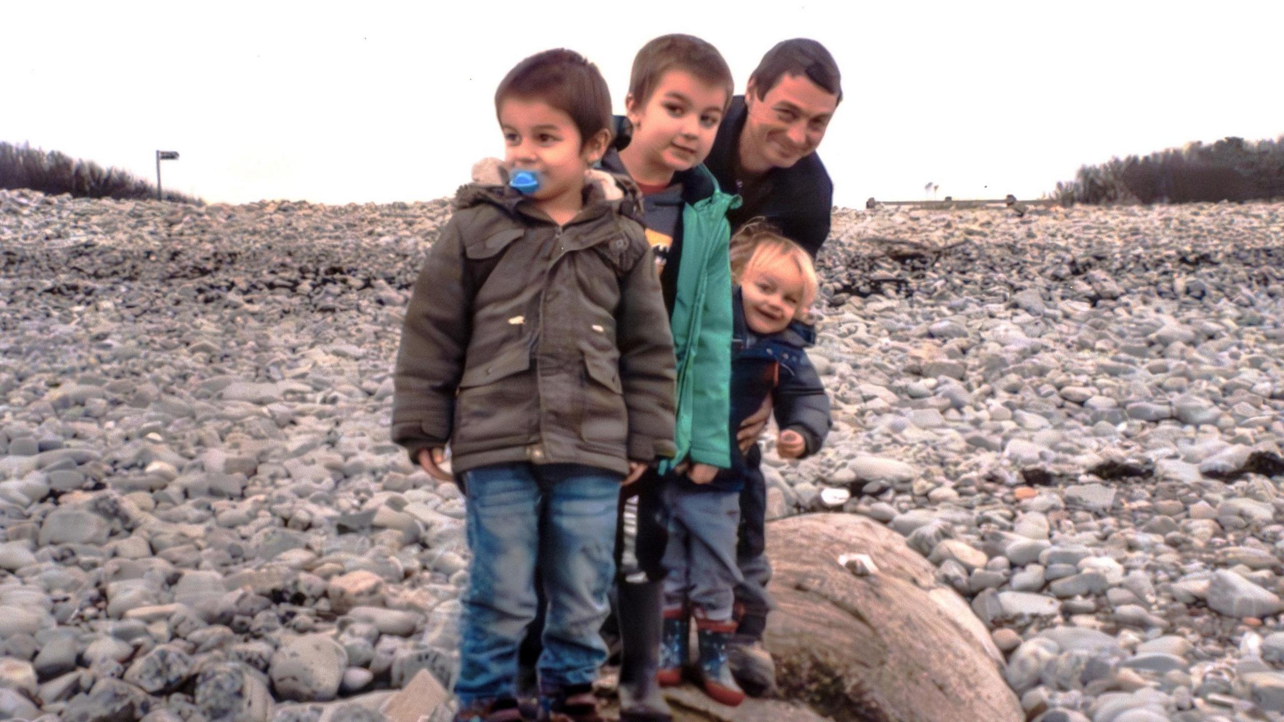 An old family photo of Sammiie's husband and her three young children on the beach. They are balancing on an old cement pipe which is coming out of the shingles on the beach. The boys are wearing jeans and coats, and the one at the front has a blue dummy in his mouth. Their father stands behind them, smiling at the camera and holding the youngest up by his waist.