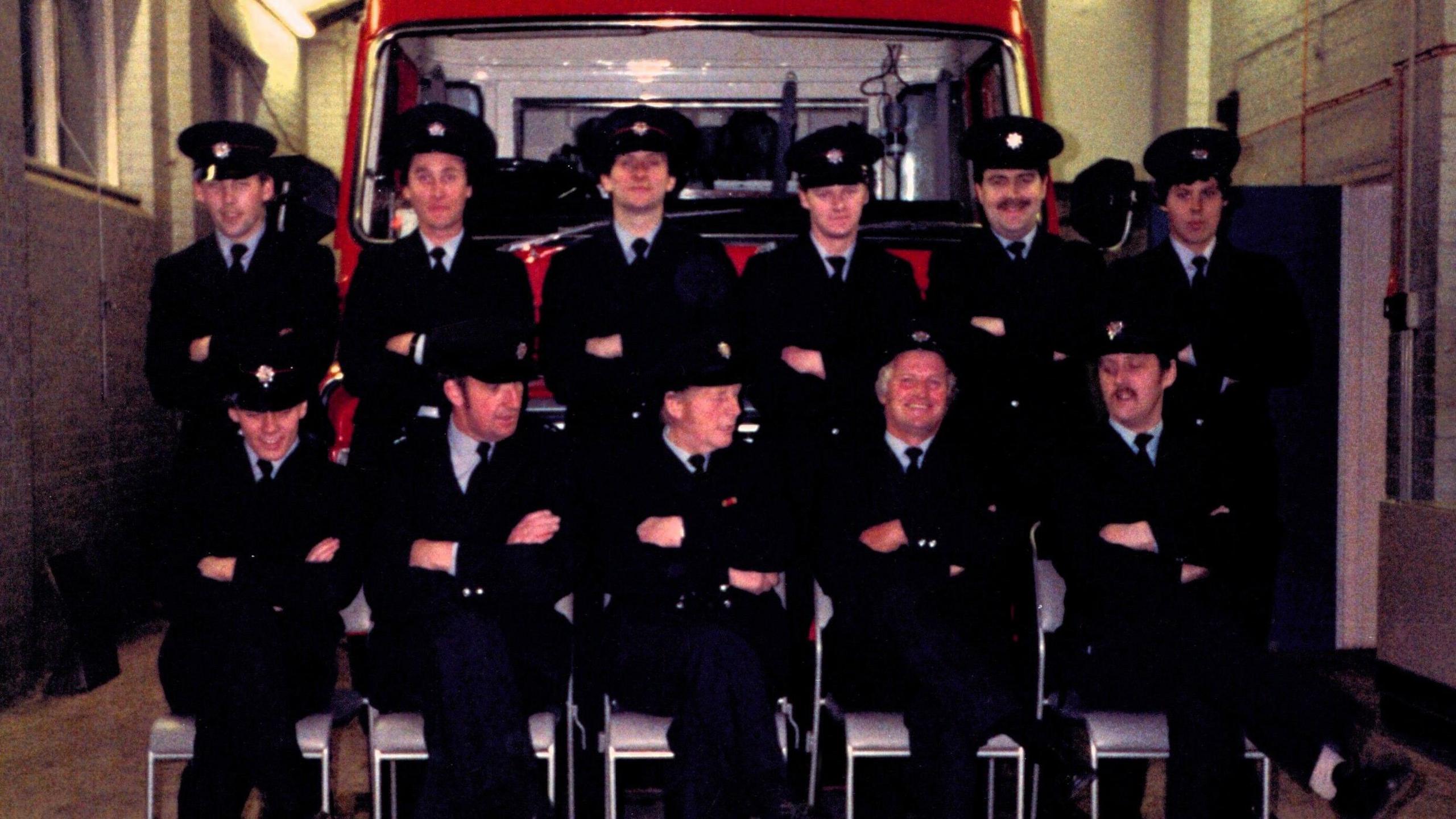 A group of 11 firefighters, including Christopher Betts, in uniform in two rows in front of a large red fire engine. The front row are seated. The back row are standing. All have their arms folded.