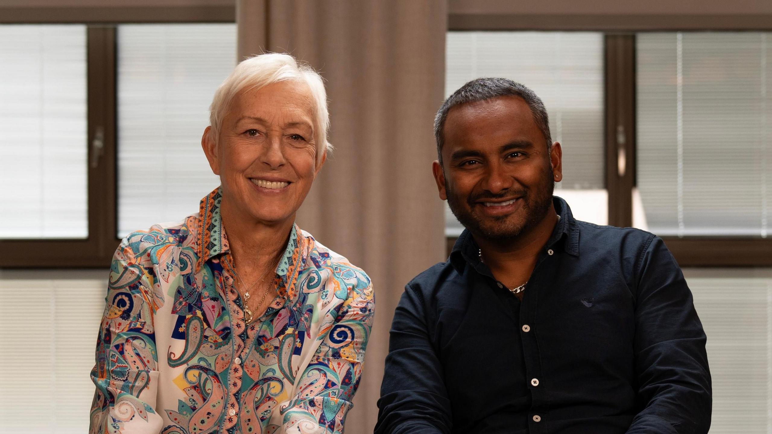 Martina Navratilova, with short white hair, wearing a colourful paisley-print blue and orange collared shirt and several gold chain necklaces. She's sitting next to Amol Rajan, who is wearing a black collared shirt. Both are smiling at the camera. The background is out of focus but there are windows and beige curtains visible.