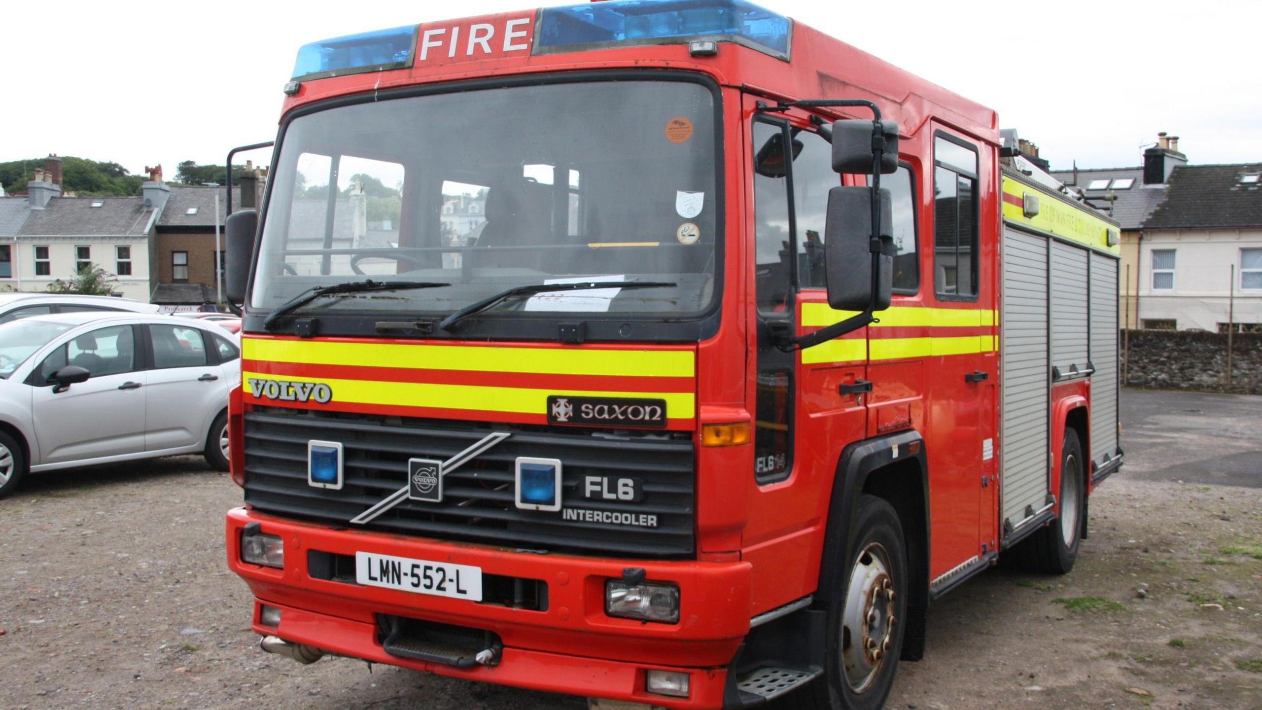 A red fire engine parked on a sandy area. There are cars parked to the left and houses in the background.