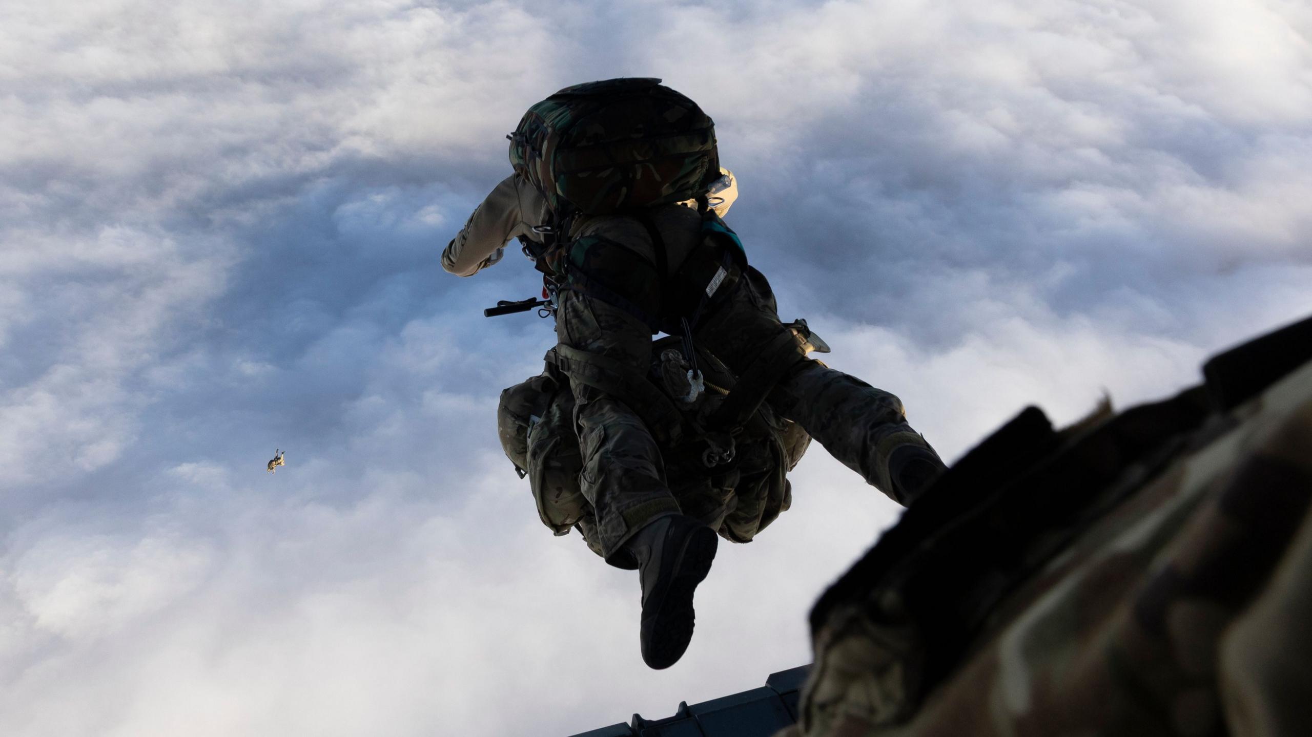 A paratrooper taking part in a parachute jump - they are seen having just left the plane and with the parachute pack on their back, with blue skies and clouds below them