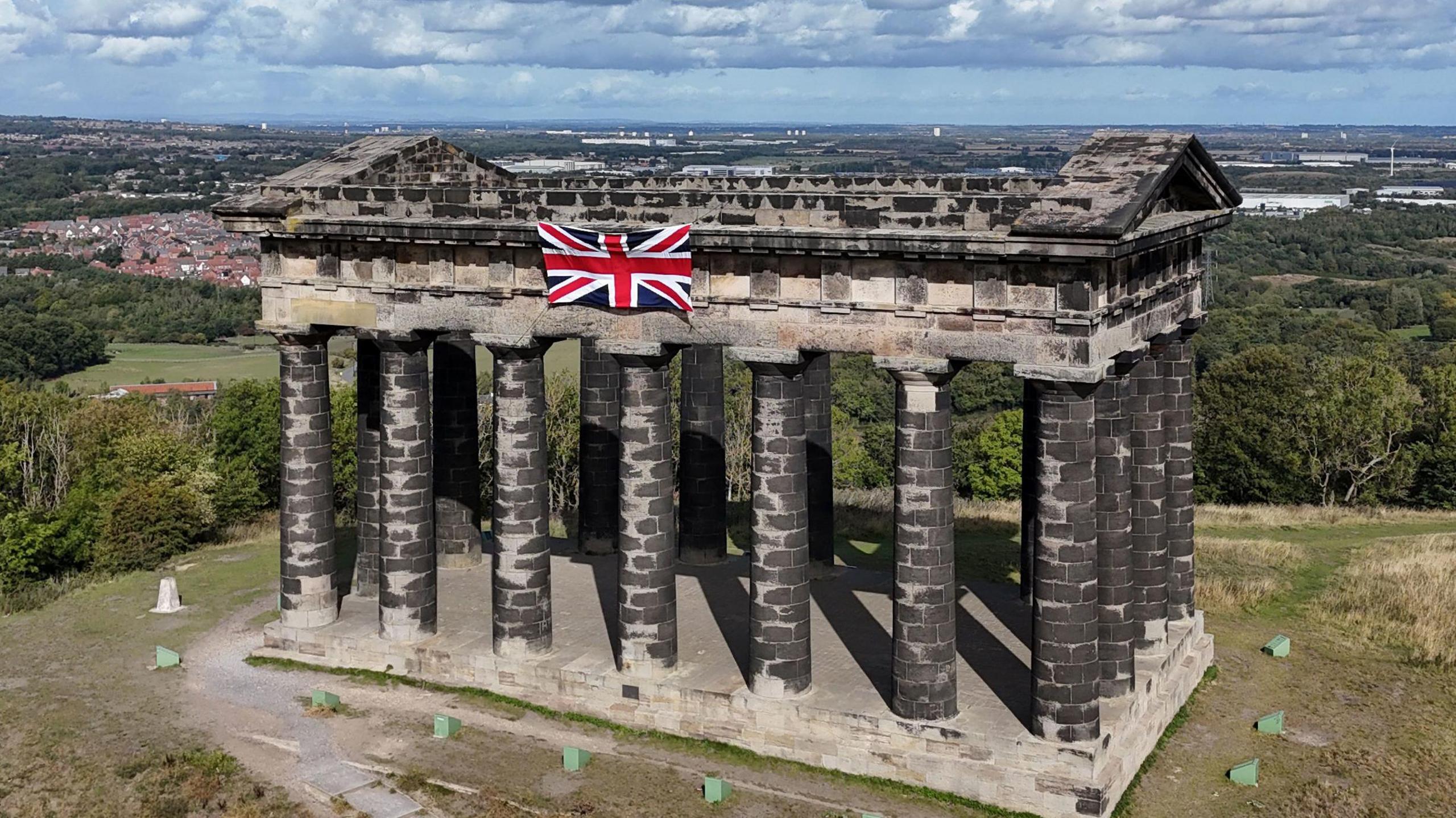 A union jack flag, flying on the Penshaw Monument on Penshaw Hill, near Sunderland. The monument consists of multiple columns made of greying stone. Countryside and urban residential areas can be seen in the distance.