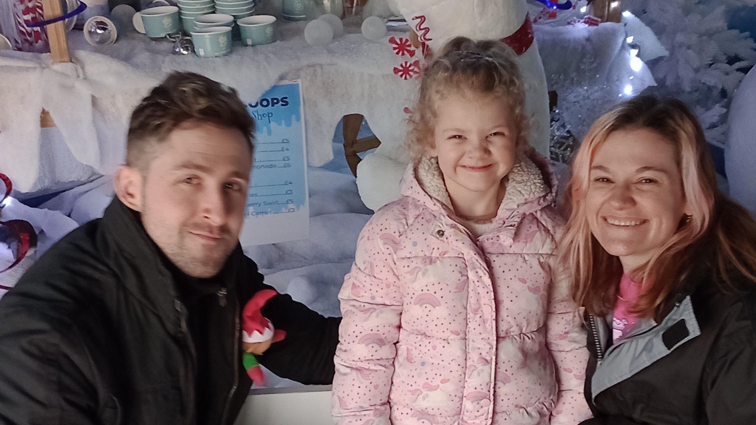 The girl and her parents from the first image. Here they are as what appears to be a christmas event. Luna, 6, wears a light pink winter coat. Both adults wear black coats. Louis has a toy elf in his breast pocket. All three people are smiling.