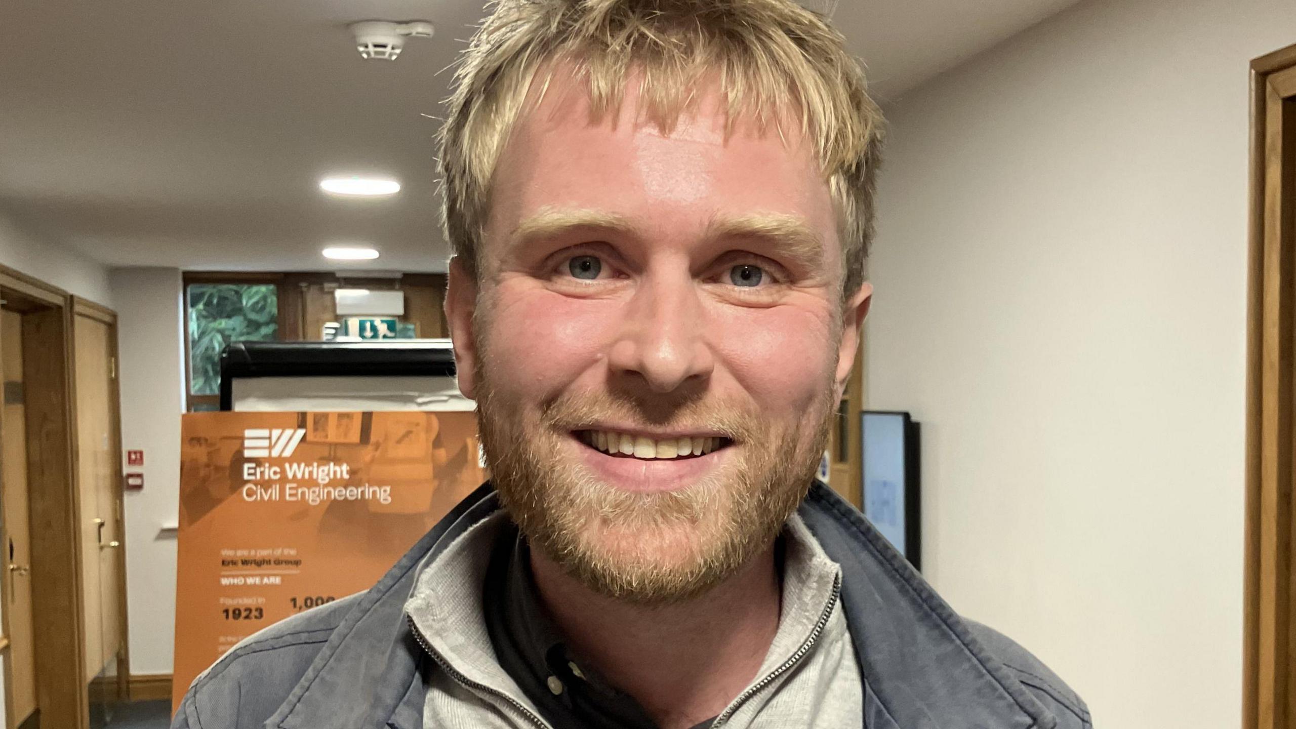 A white man with blonde hair smiling wearing a grey jumper and dark jacket.