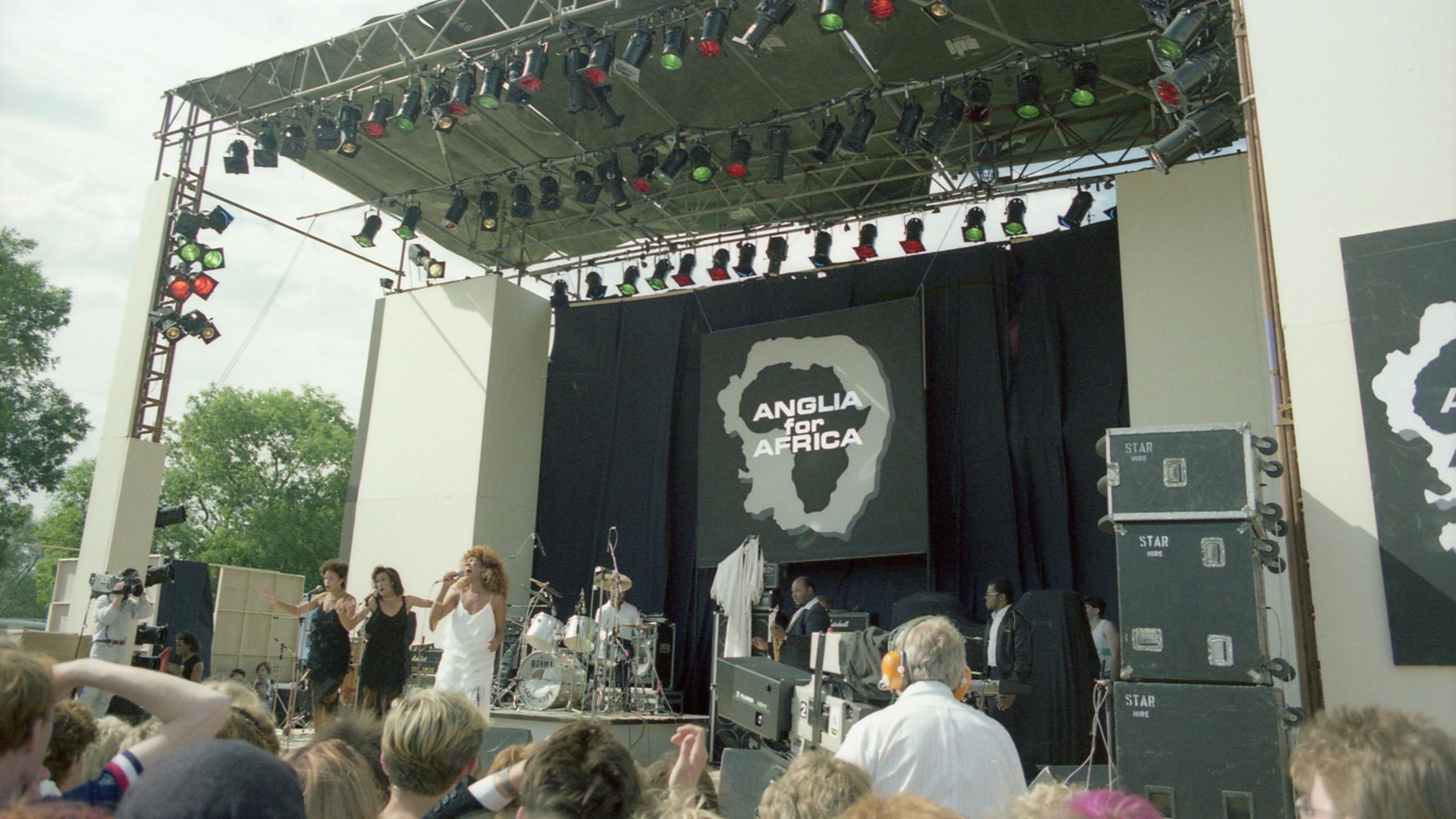 A wide shot of an outdoor concert stage seen in daylight, with a large logo at the back of the stage reading 'Anglia for Africa', over a shape of Africa inside an outline shape of Norfolk and Suffolk. Performing on the stage are three women in glamorous dresses. The backs of the heads of some of the crowd can be seen at the bottom of the frame, as can a camera operator looking into his camera as he focuses it on the group.