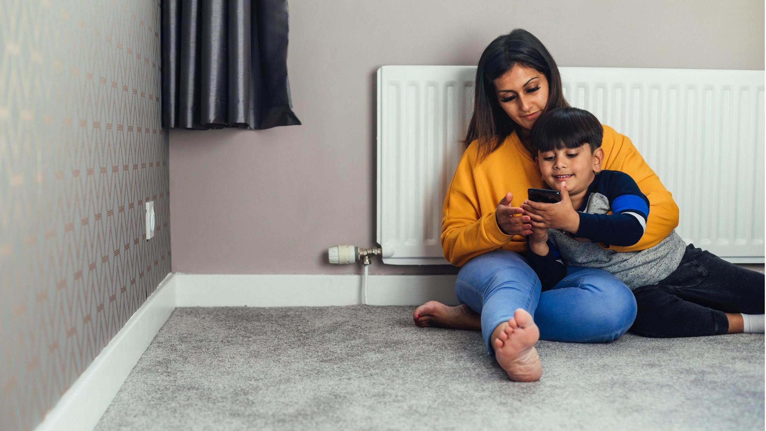 A woman and boy sitting on the floor with their back to a radiator. The boy is holding a mobile phone.