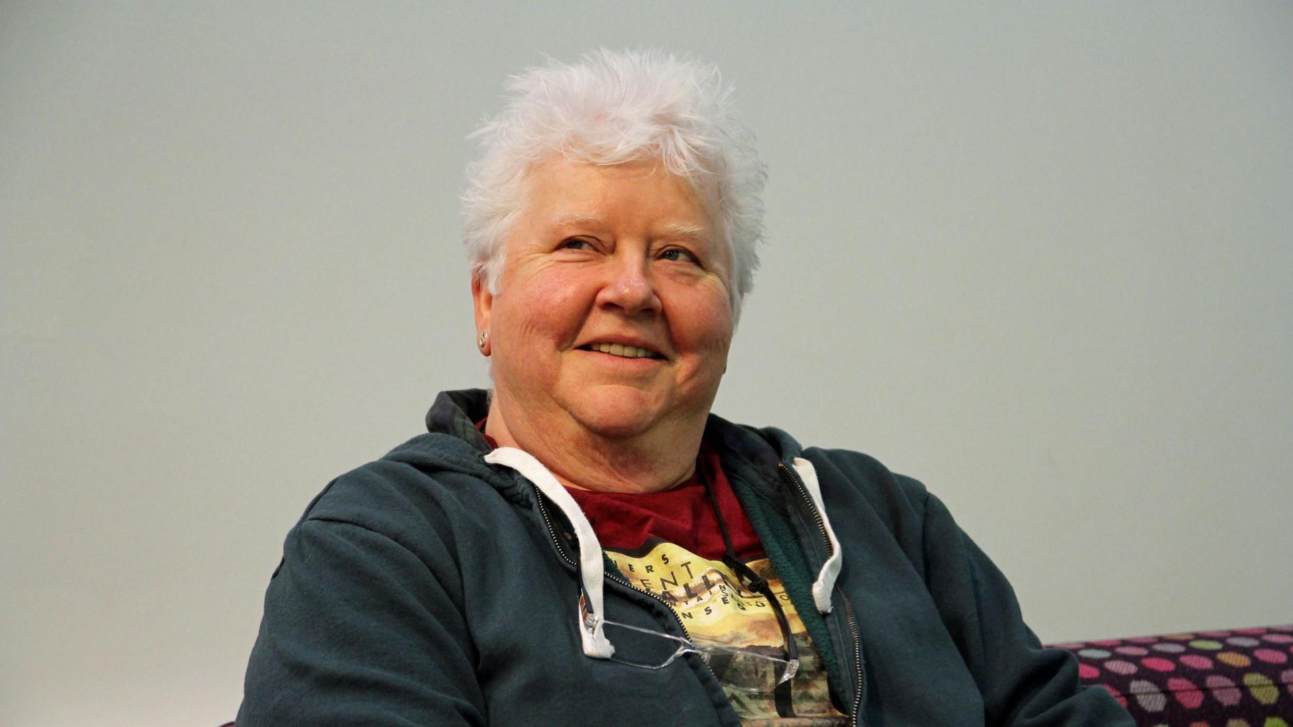 Val McDermid sitting on a couch. She has white hair, a red t-shirt and a dark hoodie or jacket on. 