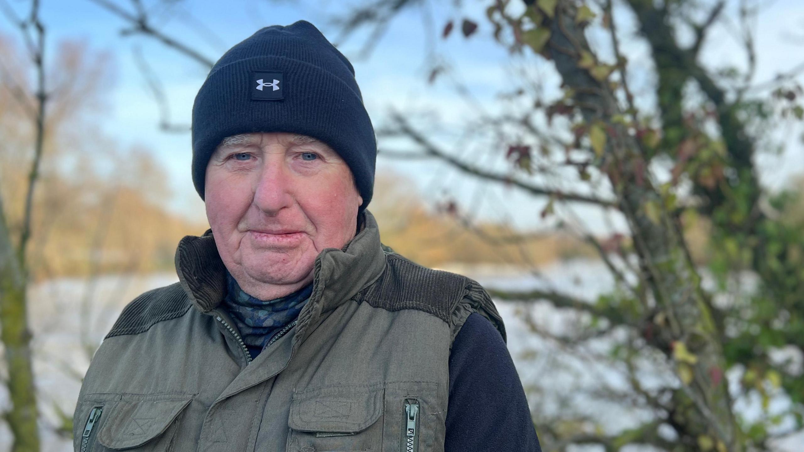 David Rennie wears a blue Under Armour beanie hat, green gillet and long sleeve blue jumper. He is stood on the footbridge and there are trees, with no leaves, behind him. Through the gaps in the trees the Great River Ouse can be seen.