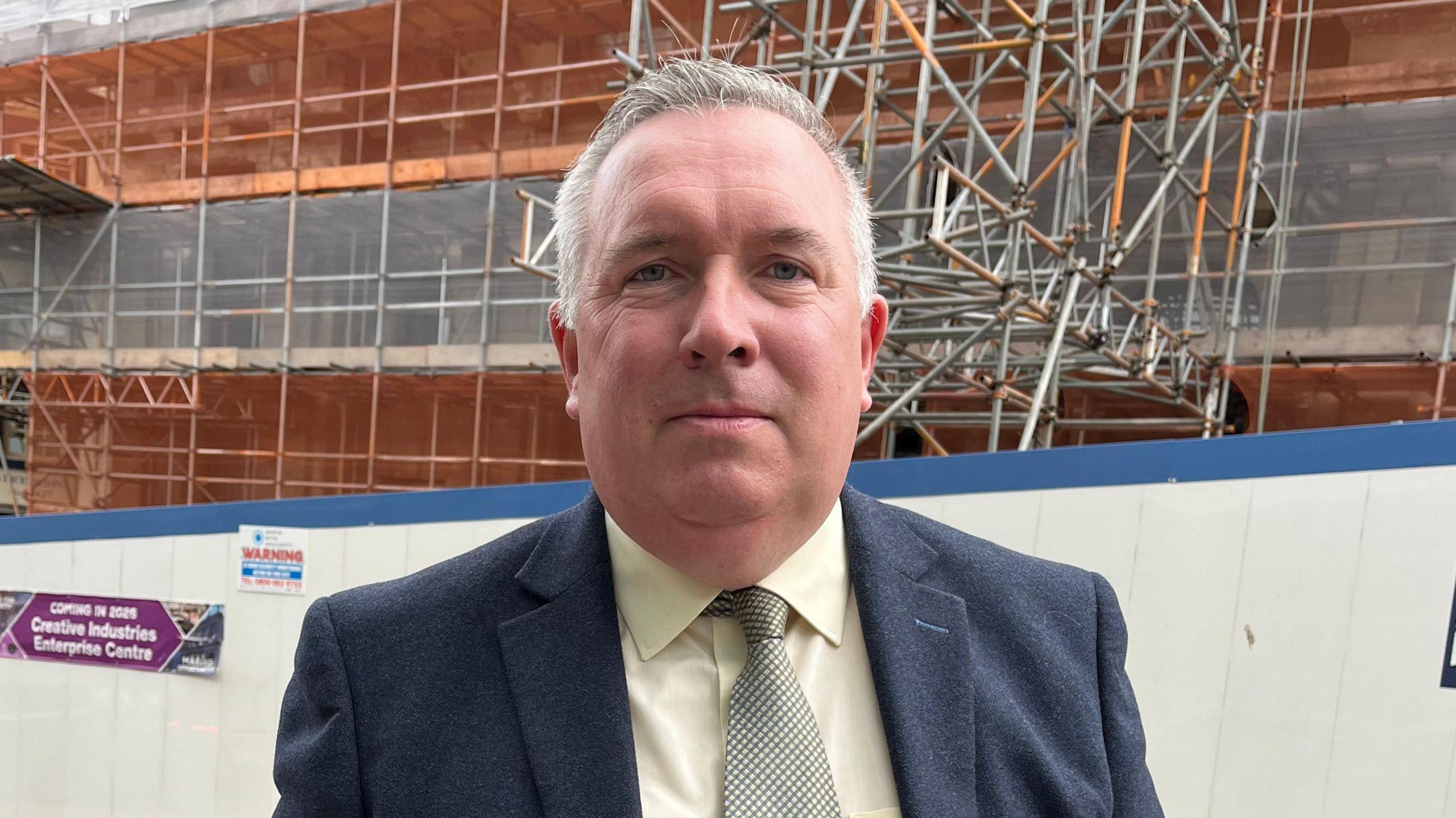 Deputy leader of the council, Adrian Andrew wearing a blue suit stood in front of scaffolding. He has short greying hair and wears a dark blue suit over a light green shirt and patterned green tie.