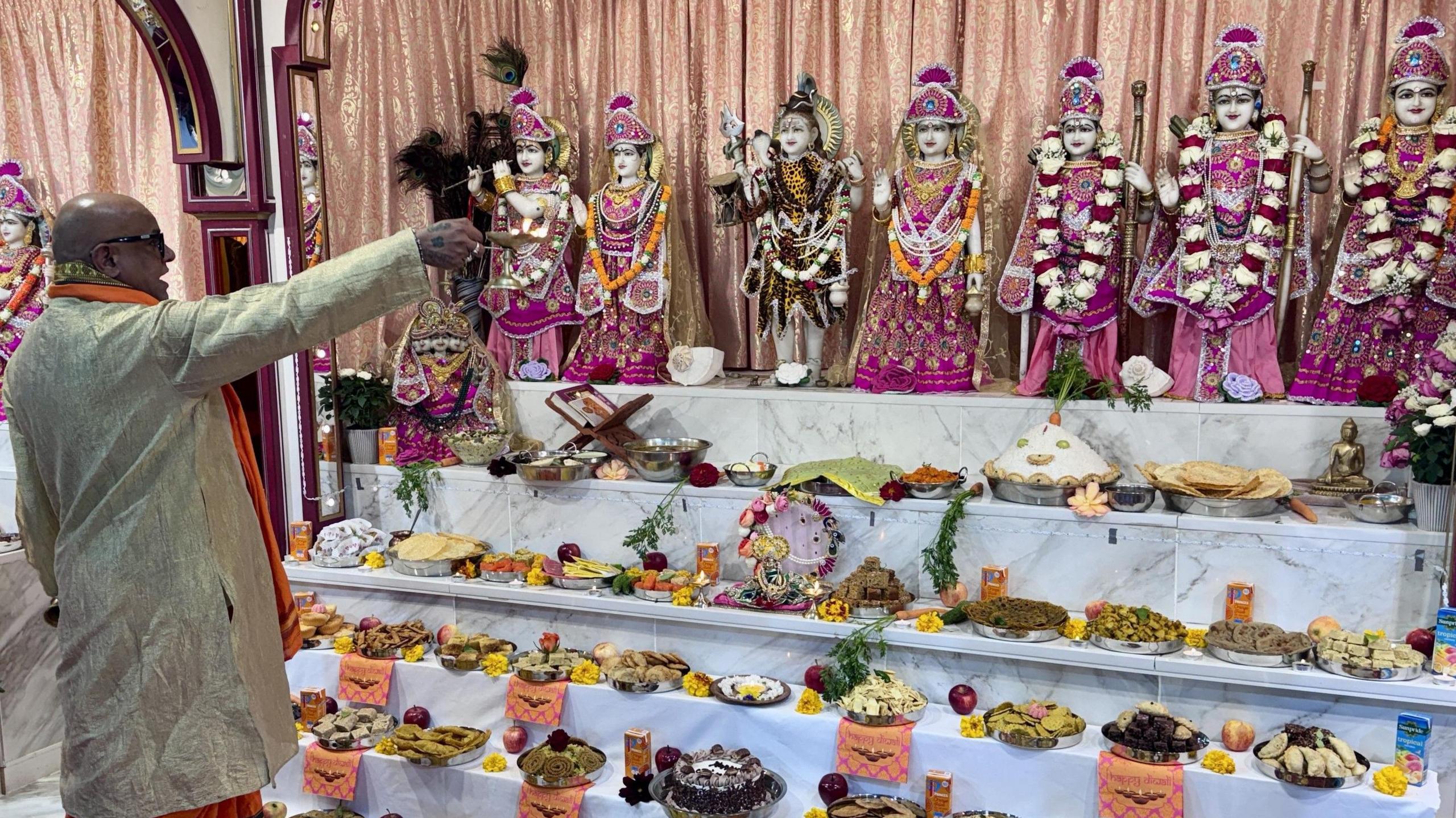 A Hindu priest prays to deities in the Main Sanctum of the Hindu Temple. He is holding a candle holder with a lit candle. He is stood in front of a colourful display showing lots of dishes of food which are being offered to the gods.