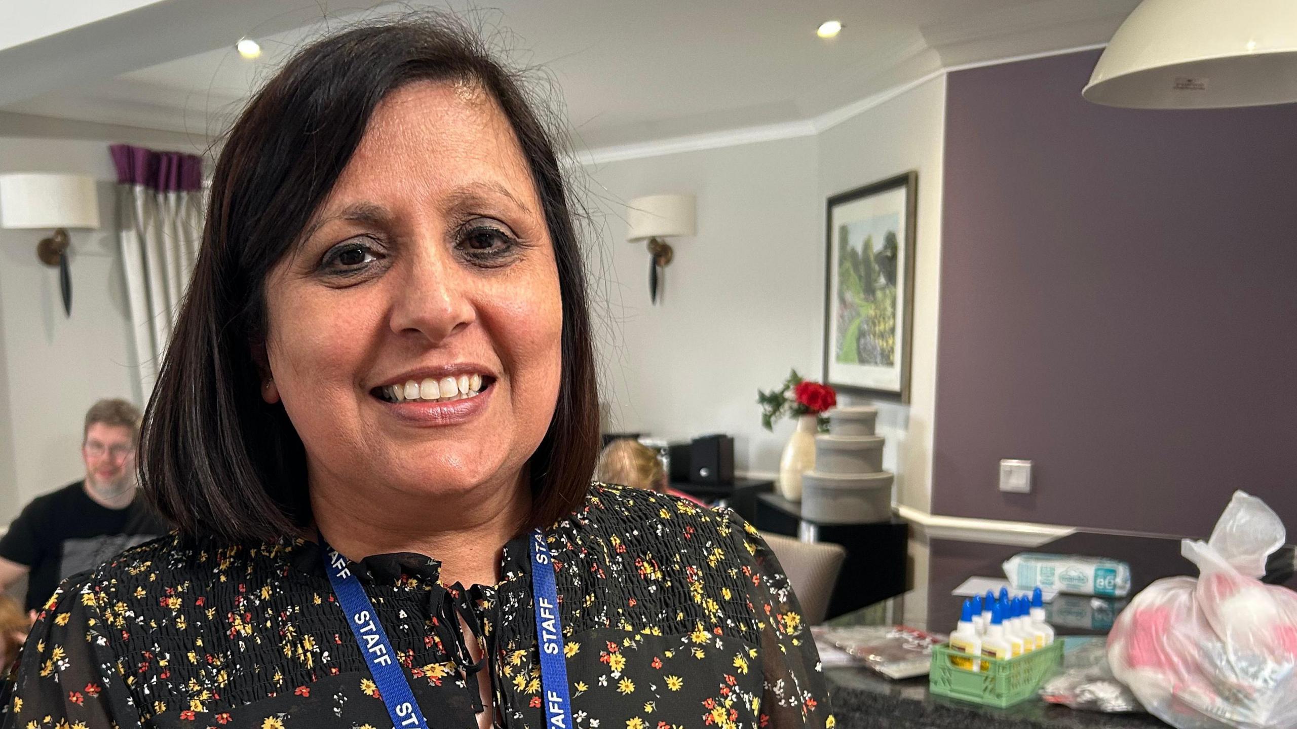 Kuldeep Lidder, who has shoulder-length black hair and is wearing a black top flecked with yellow, white and red flowers. She is standing in a room with a table to her left with art supplies on it