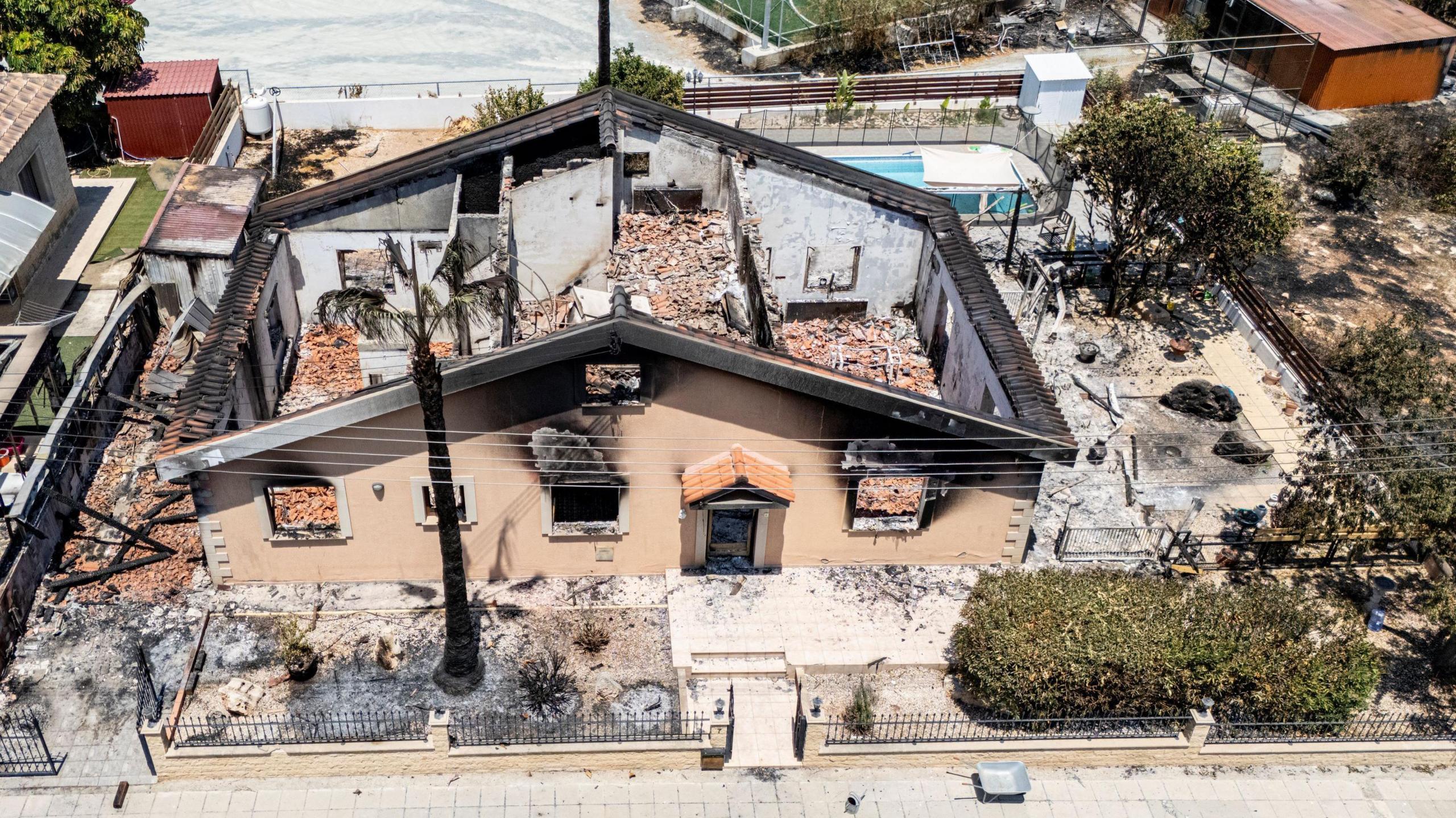 Birdseye view of the ruins which remain of the family home in Sinou, Cyprus. Burnt black grass can be seen outside of their fence on the left. The home is left without a roof, with only the house walls remaining among the rubble. 
