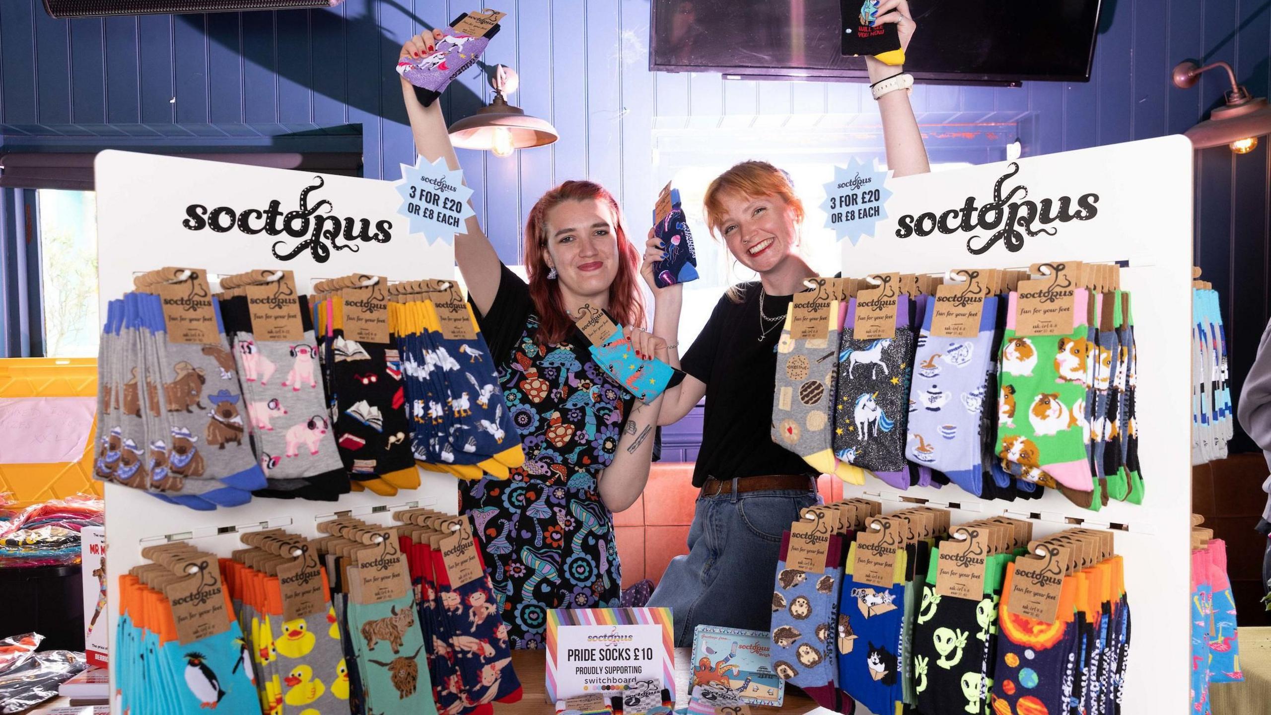 Two women stand in a shop surrounded by unique, brightly coloured socks. They both have red hair.
