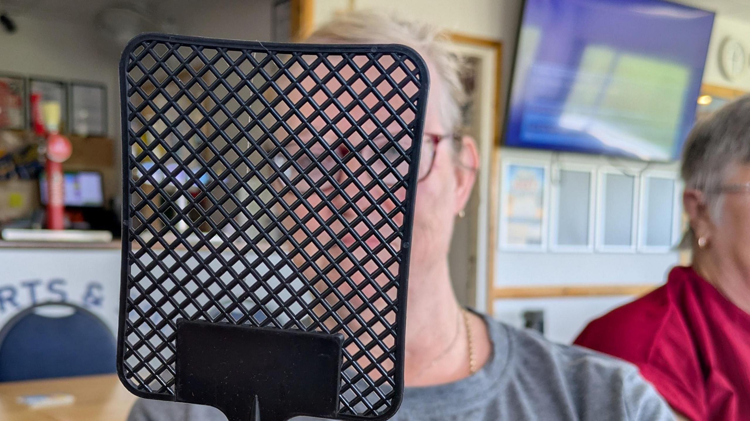 A woman with short grey hair and gold earrings wearing a grey T-shirt sitting in a sports clubhouse with a black fly swatter in front of her face.