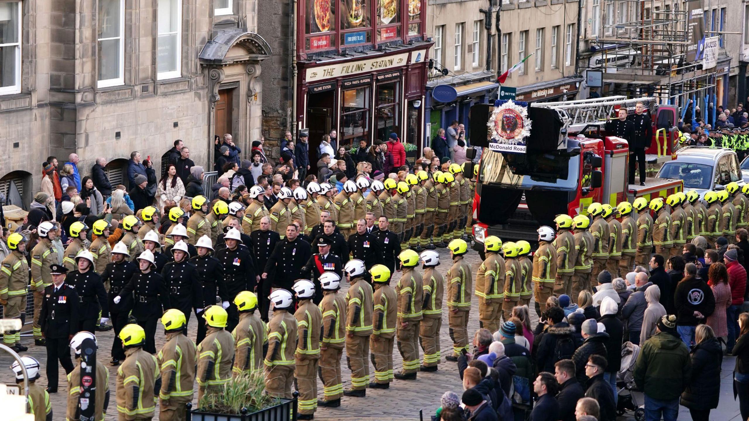 The funeral procession for Barry Martin in Edinburgh - a fire engine drives up the street carrying the coffin, proceeded by various firefighters dressed in black. Uniformed firefighters line up on either side, with rows of locals standing behind them watching.