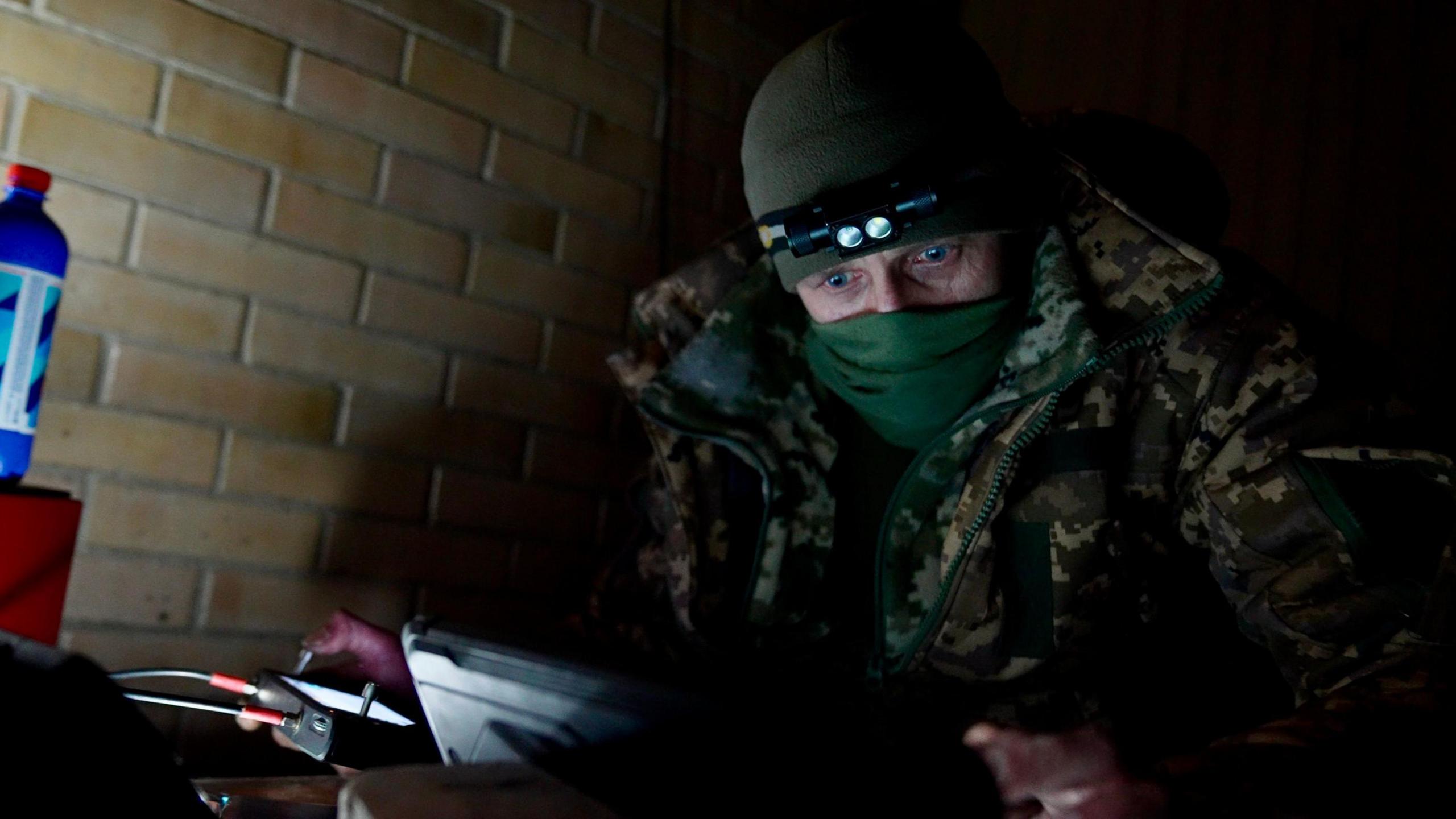 Soldier with mask covering his mouth and nose, in a hat with head torch, looks intensely at a screen whilst holding a controller