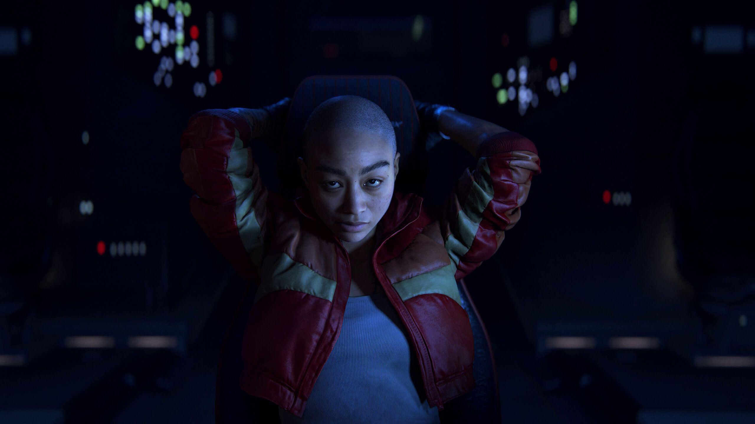 A woman with a shaved head wearing a red leather jacket sits in a darkened room with both hands placed behind her head. She's illuminated by lights from nearby control panels and screens.