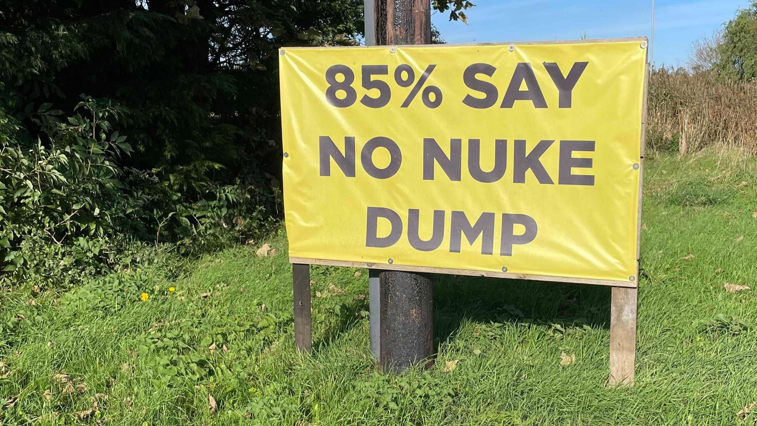 A yellow banner is stretched between two poles in front of a telegraph pole on a grassy stretch of land. Large, black capital letters read: "85% say no nuke dump". 