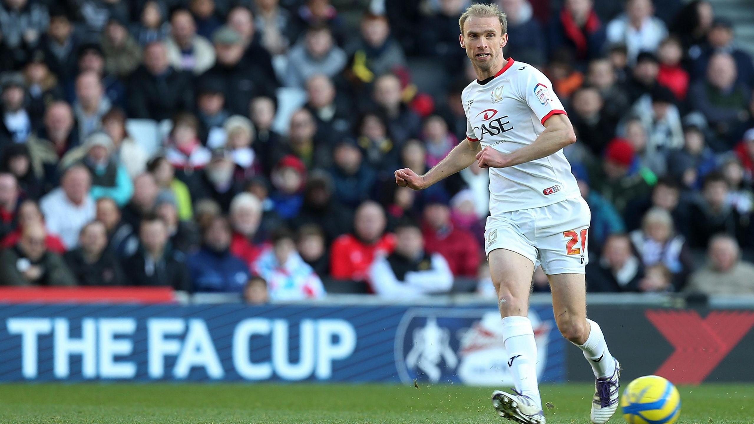 Chadwick, in a full white MK Dons kit, passing the ball.