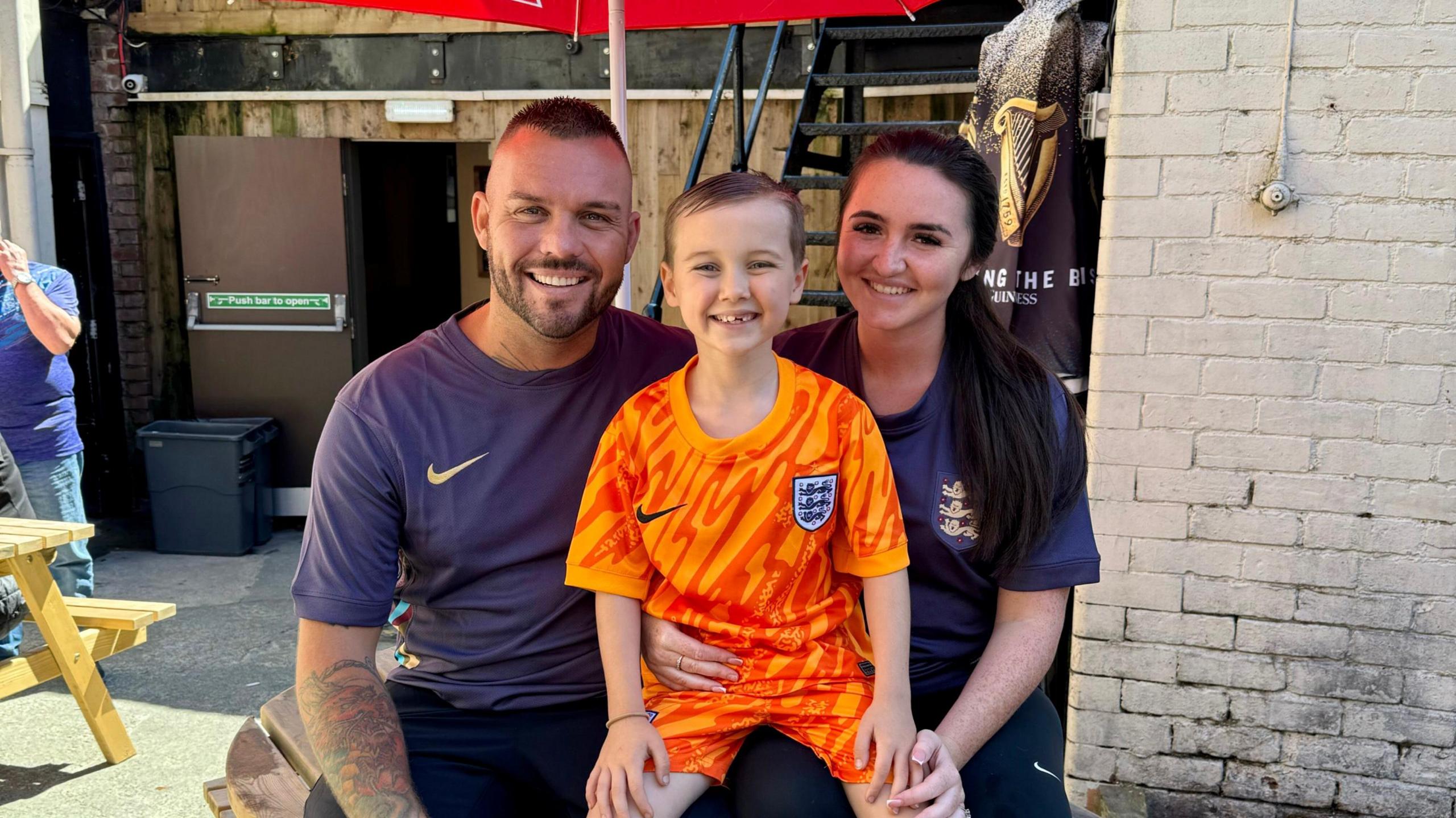 Church Inn landlady Steph Savage sits on a picnic table outside her pub along with her husband and son, each of them wearing England shirts