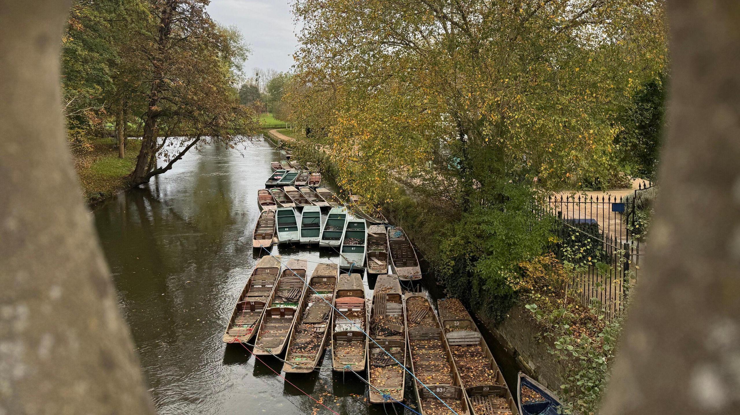 A view taken from Magdalen bridge in Oxford of punts on the River Cherwell.  Two of the bridge's stone balusters frame the edges of the picture. The punts on the water are filled with autumn leaves and are tied to the riverside. Metal railings line the waters edge.