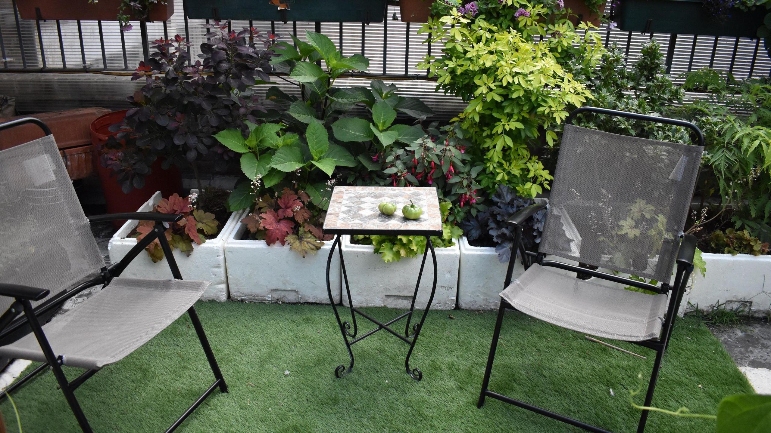 A set of garden furniture, with two deck chairs and a small table, on a patch of artificial grass. These are placed in front of a set of white planters, filled with a variety of leafy plants.