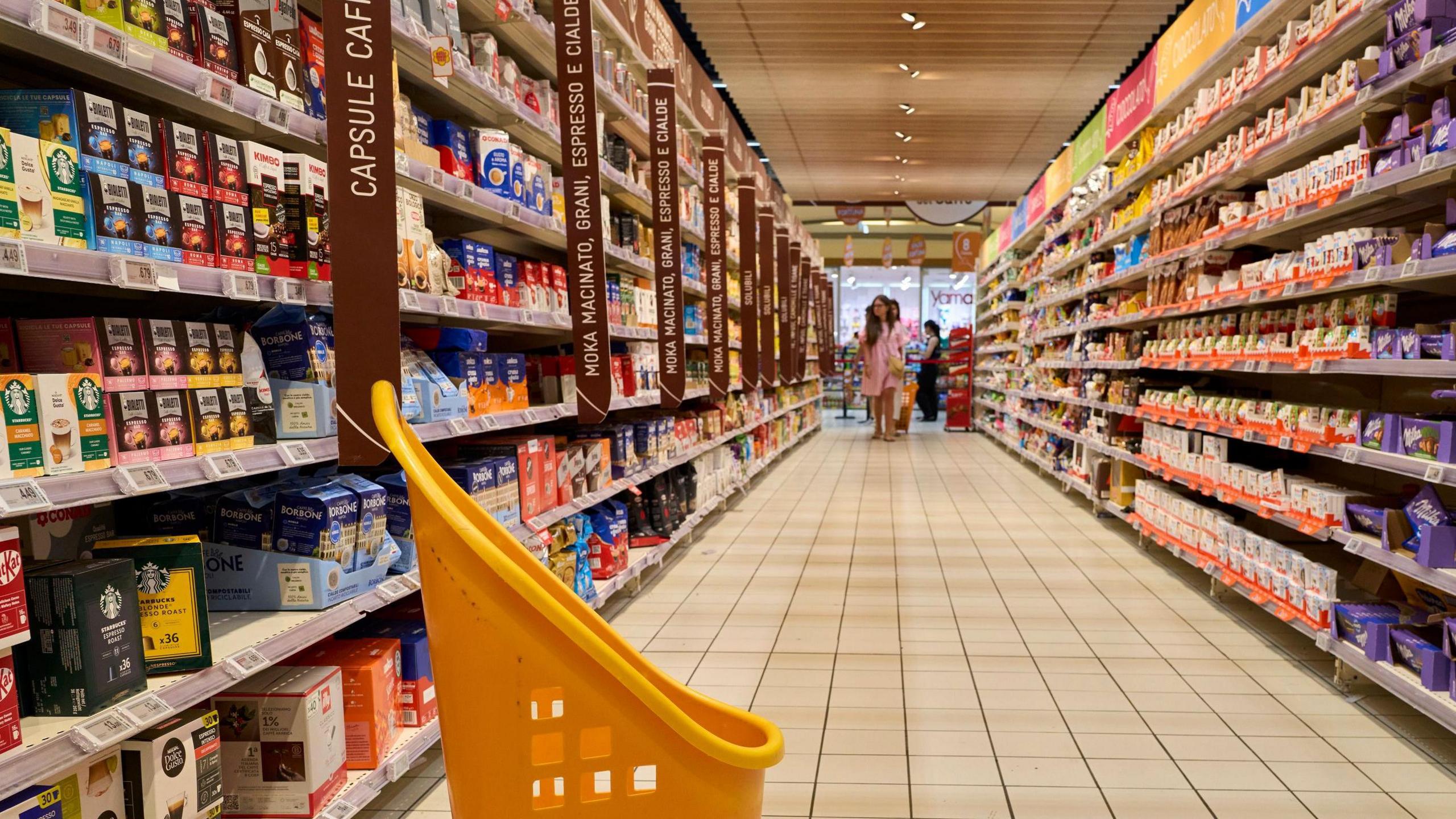 A supermarket aisle with shelves stocked with various coffee products on the left and other grocery items on the right. The aisle is well-lit, and a yellow shopping cart is visible in the foreground. In the background, two people are walking down the aisle towards the camera.