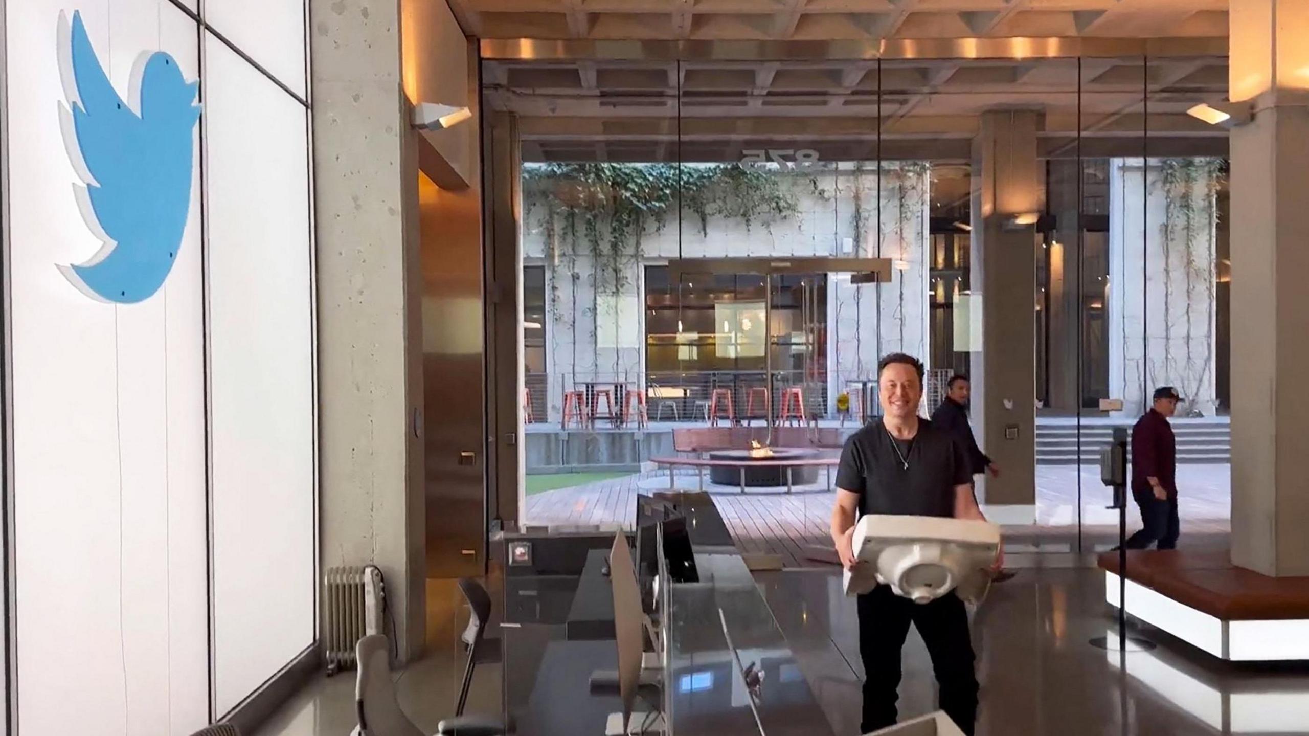 Elon Musk carries a sink into the reception of the headquarters of X, then known as Twitter, on the wall is the old Twitter logo.