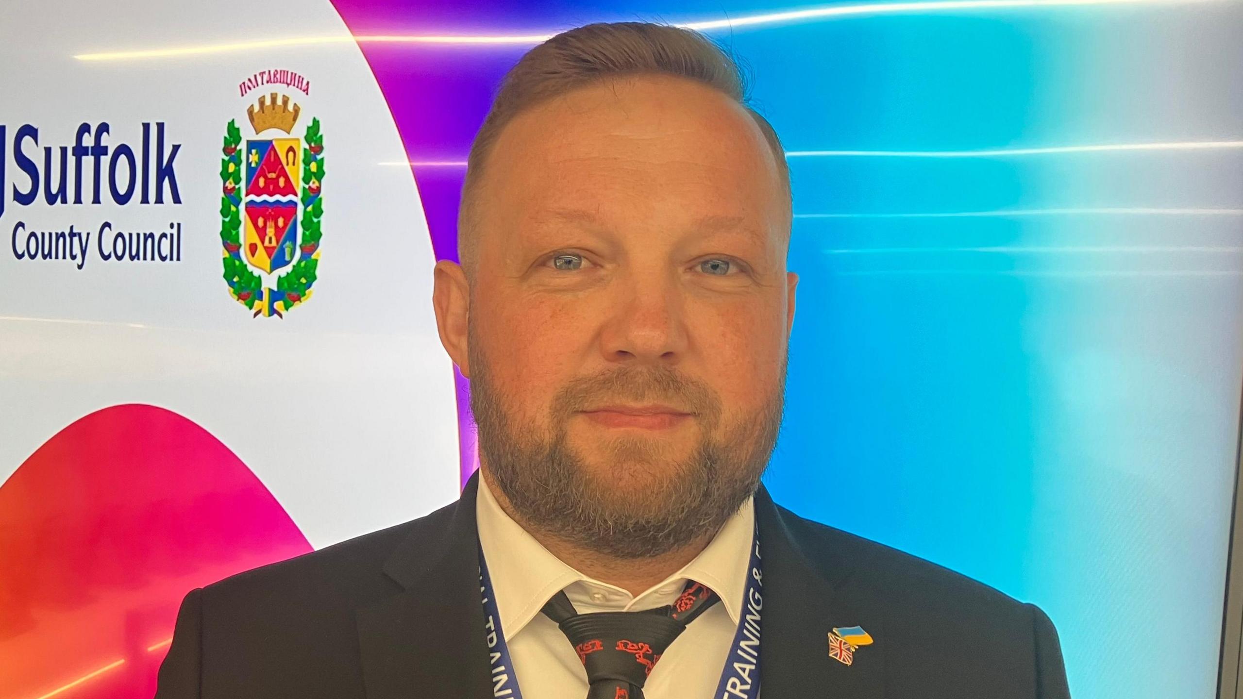 Volodymyr Kohut looks at the camera with a light smile. He has short brown hair and is wering a black suit jacket and a white shirt and blue and red tie. He is standing in front of a screen that says "Suffolk County Council".