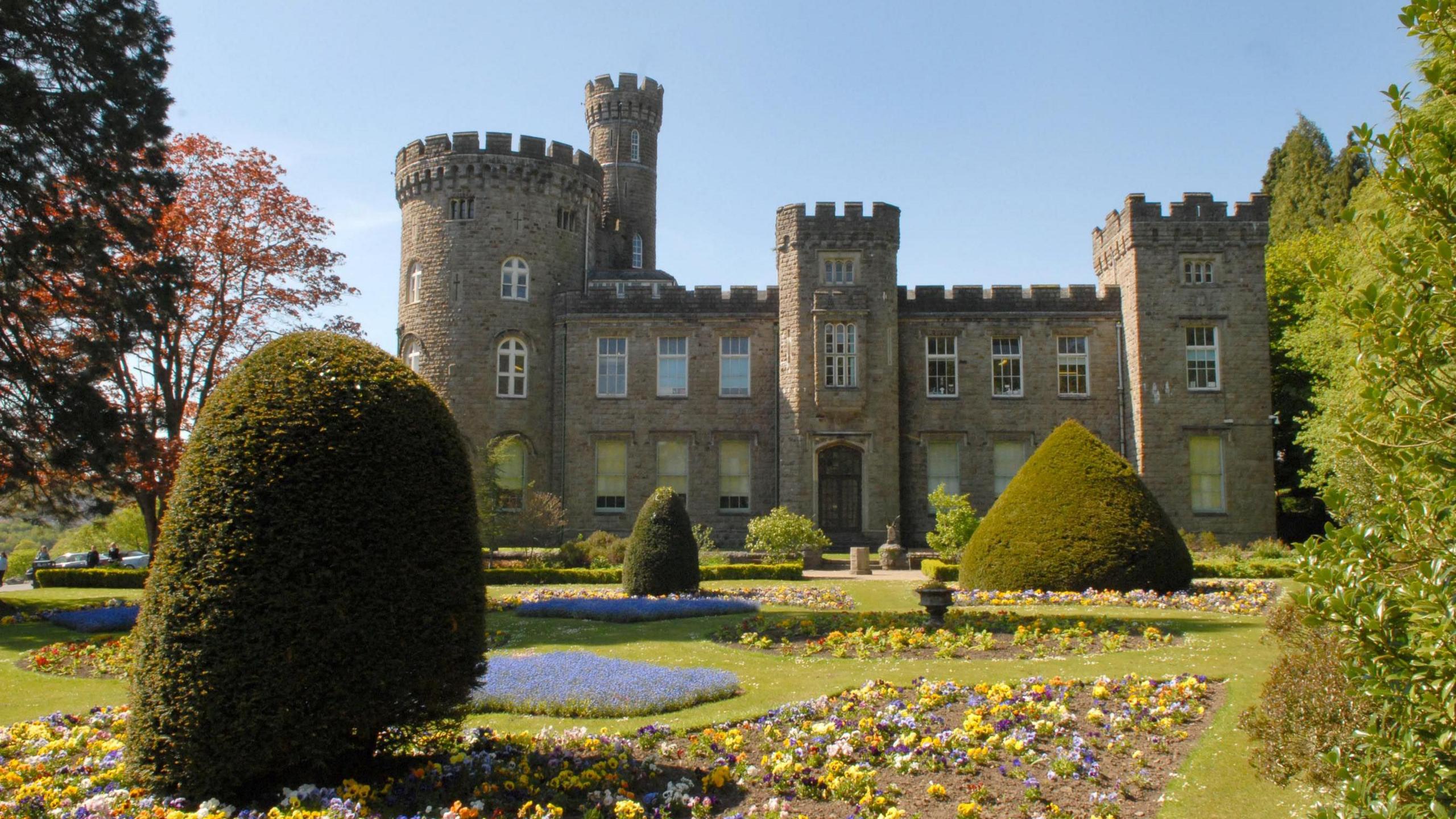 An exterior view of Cyfarthfa Castle in Merthyr Tydfil. It's front right turret is square, whereas the central and left and rounded and there is a tall, circular tower to the rear left of the building. In front there are well-manicured gardens with shaped hedges and patches of flowers with predominantly yellow, purple and blue flowers.