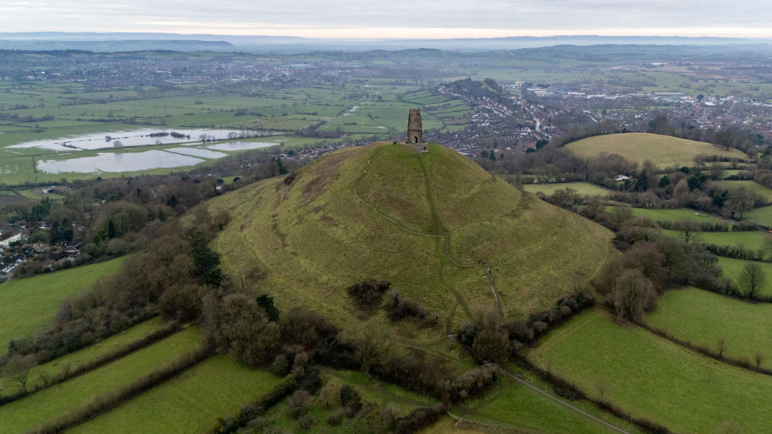 A view of Glastonbury Tor taken on a clear day from the air. The Tor is in the middle distance while large sections of Somerset countryside are also visible in the background, with many green fields and hedgerows