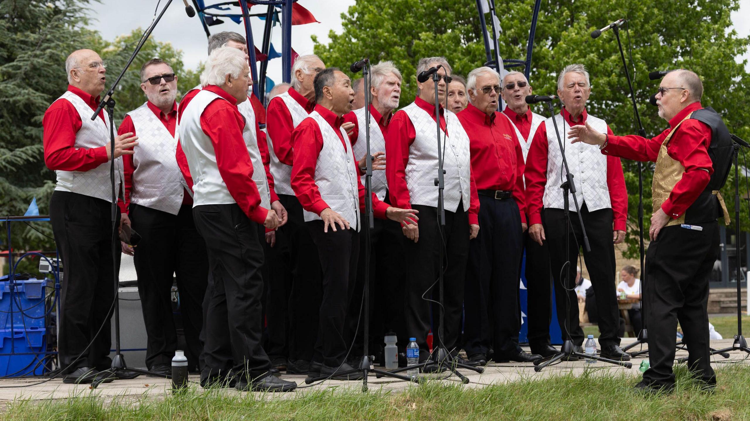 A male-voice choir wearing white waistcoats and red shirts sing whilst being conducted by a man wearing a gold an black waistcoat.