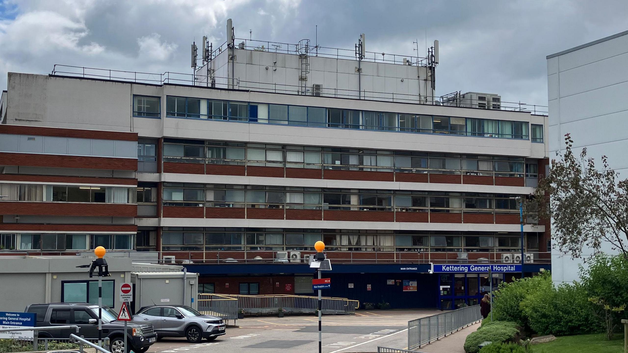 An outside view of Kettering General Hospital building. A multi-storey building with a blue entranceway with "Kettering General Hospital" is large letters above the door. There are two cars parked outside the hospital. 