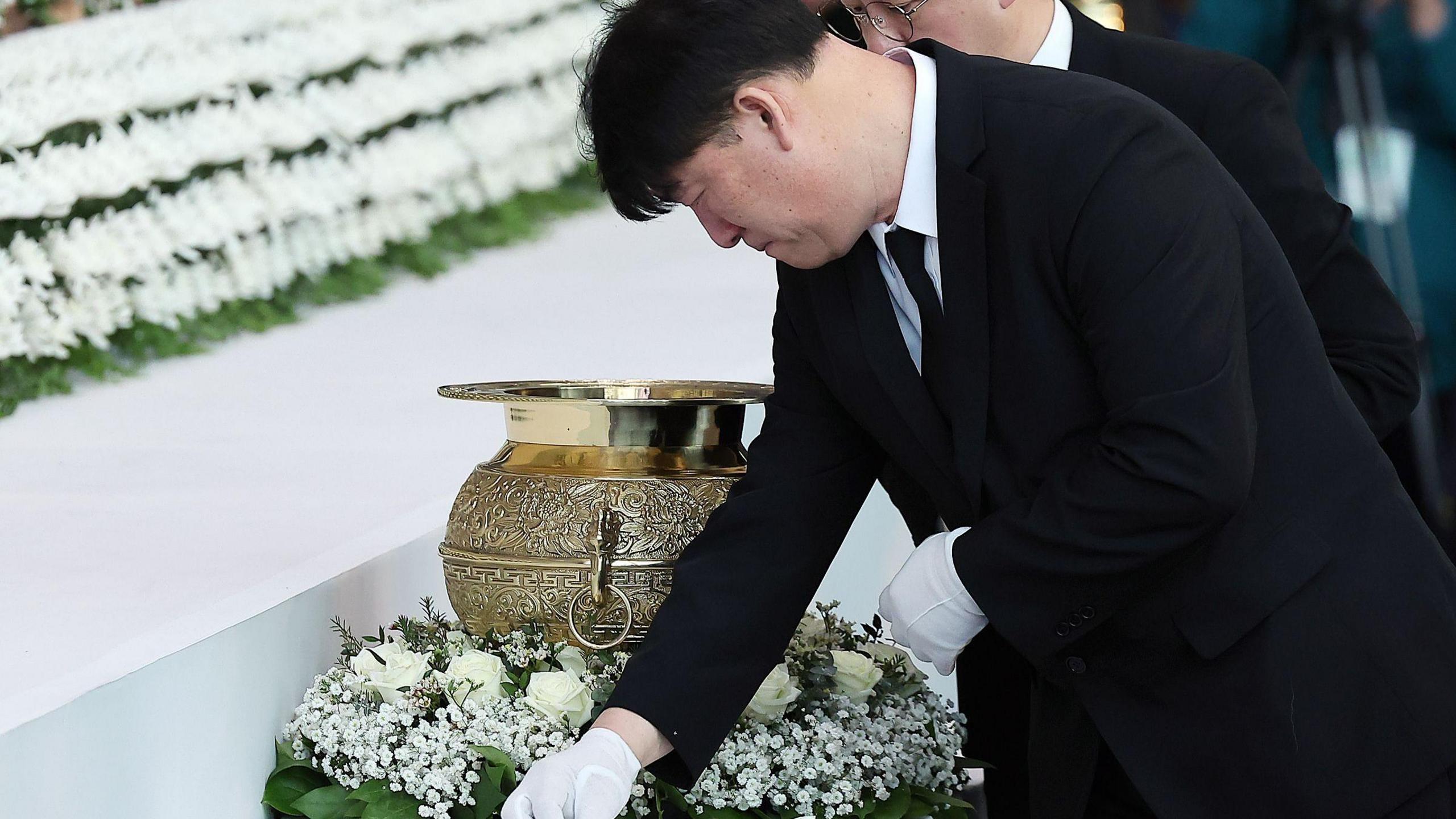 A man in a black suit and white gloves lays a flower down on a table next to a wreath and a golden urn