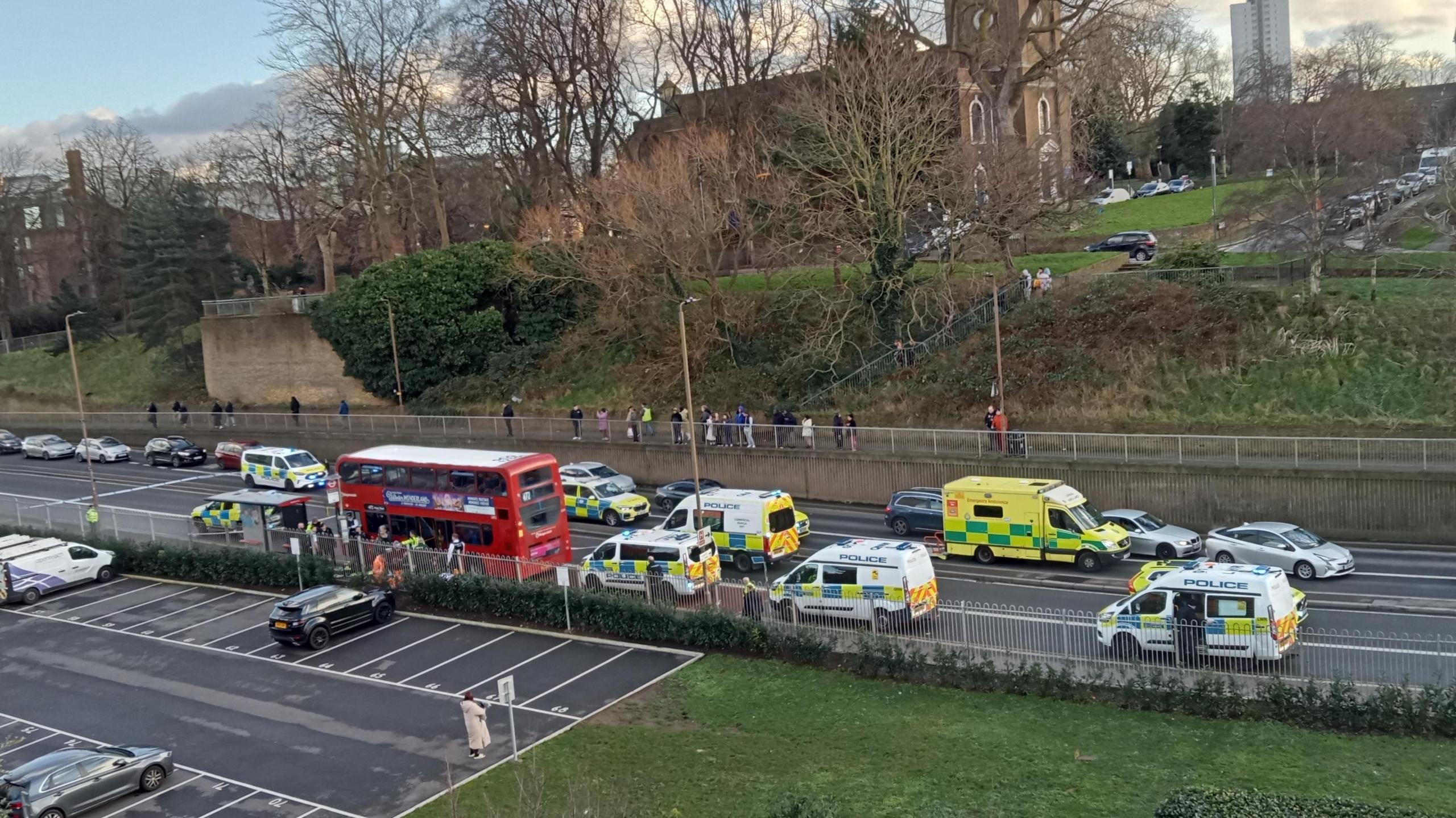 Emergency vehicles, including police cars, ambulances, and a fire engine, are parked along a busy road next to a red double-decker bus. Pedestrians observe from a pathway on an adjacent footpath.