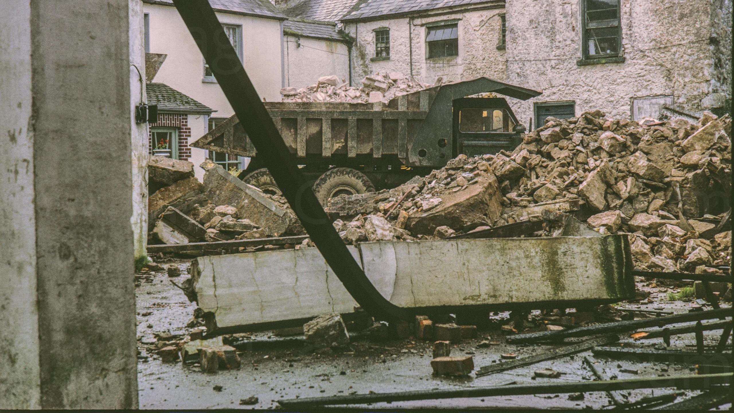 A digitalised photo from a film of a truck filled with bricks and surrounded by rubble. A lorry full of rubble stands in a yard of demolition material surrounded by a house and its out buildings. Probably at the redevelopment of Barrack Street.