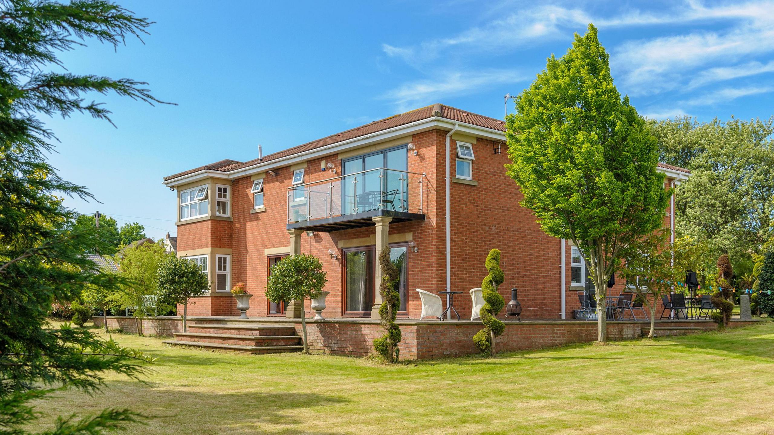 Red Gables. It is a two-storey brown-brick house with a balcony on the second floor and a seating area outside. It is surrounded by trees and other plants. 